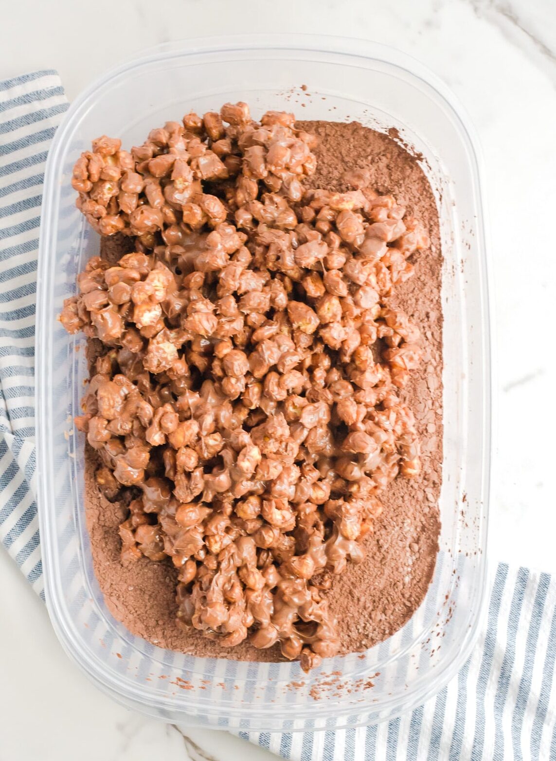 A large plastic container filled with chocolate covered Corn Pops on top of cocoa powder.