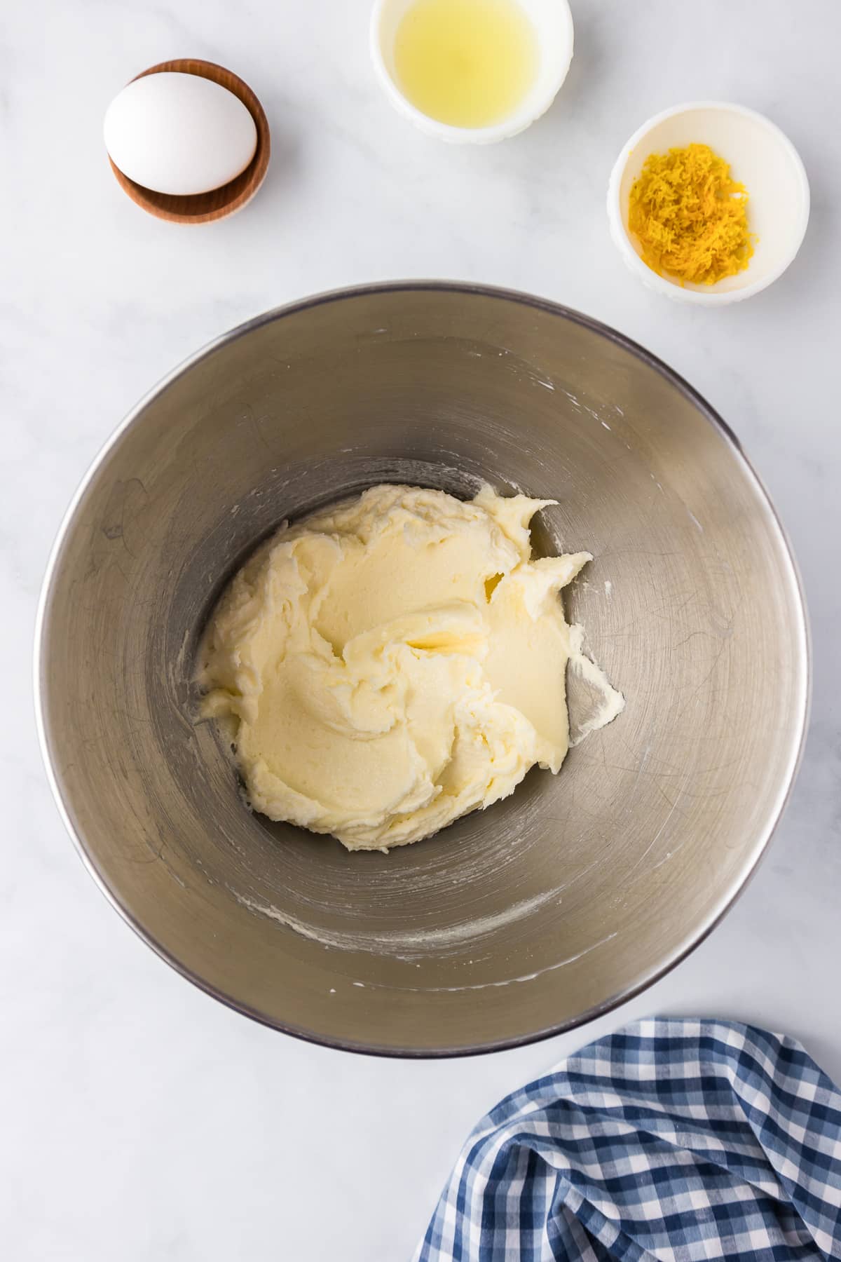A mixing bowl with butter and sugar creamed together, with lemon juice, lemon zest and an egg on the counter nearby.