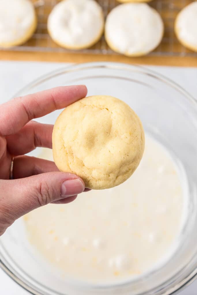 A person holding a lemon cookie over a glass bowl full of lemon glaze.