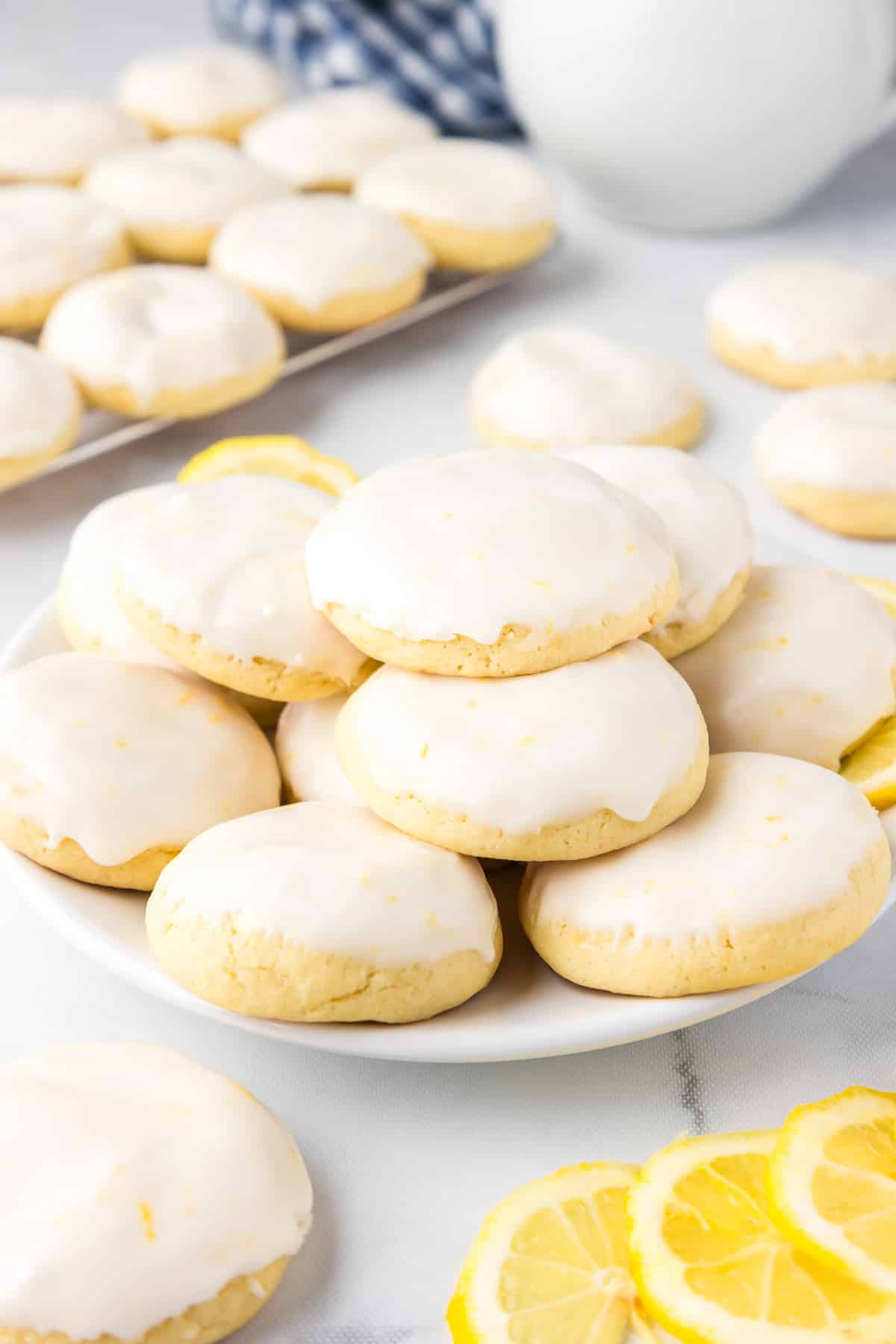 Lemon cookies with glaze on a plate with lemon slices and more cookies on the counter in the background.