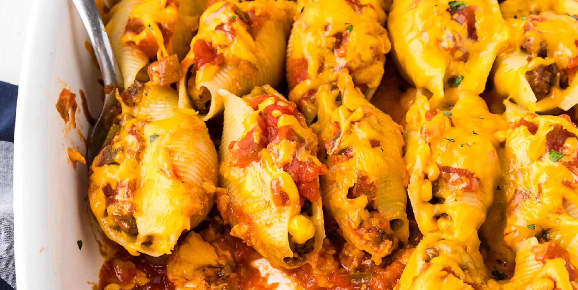 A wide view of a row of cheesy Mexican stuffed shells in a casserole dish with a spoon scooping one3.