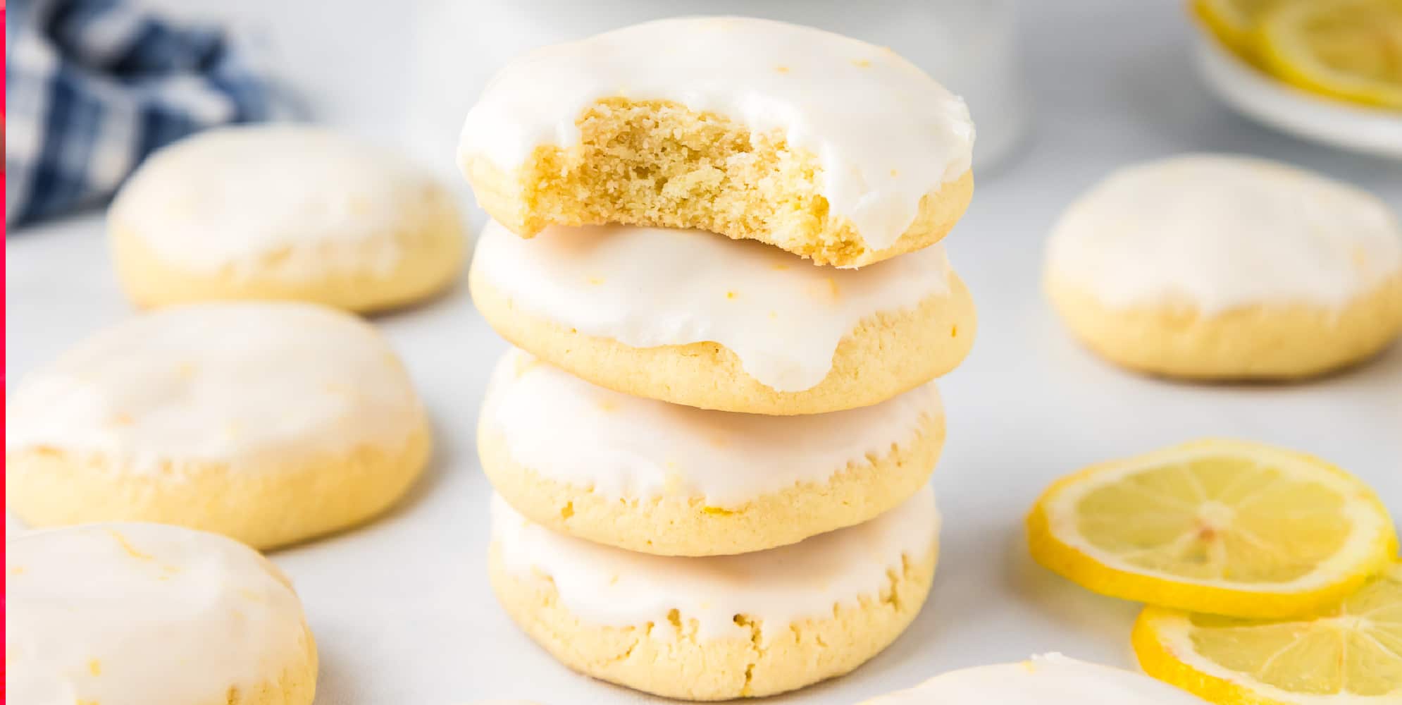 A stack of lemon cookies with a bite taken out of the top cookie with more cookies and slices of lemon nearby on the counter.