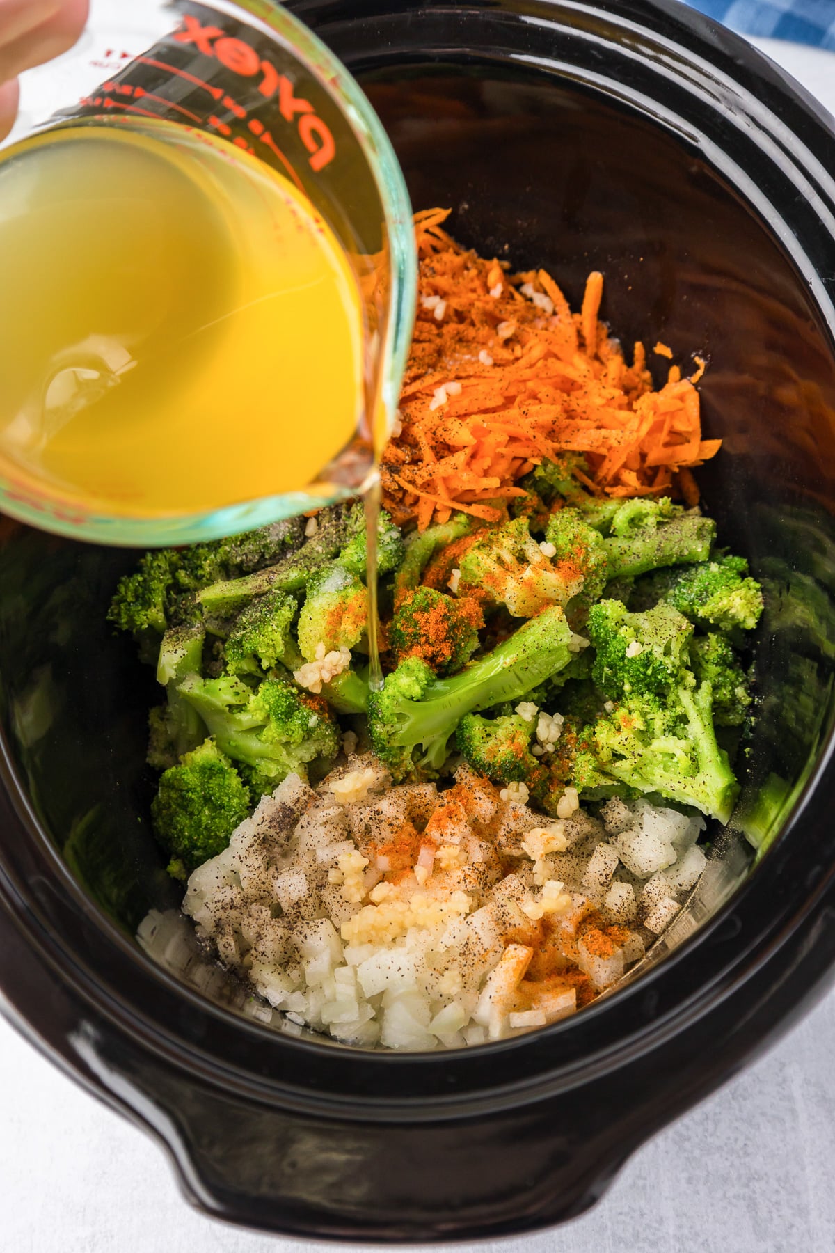 A person pouring broth into a crock pot with broccoli, carrots and other vegetables and spices for broccoli cheese soup.