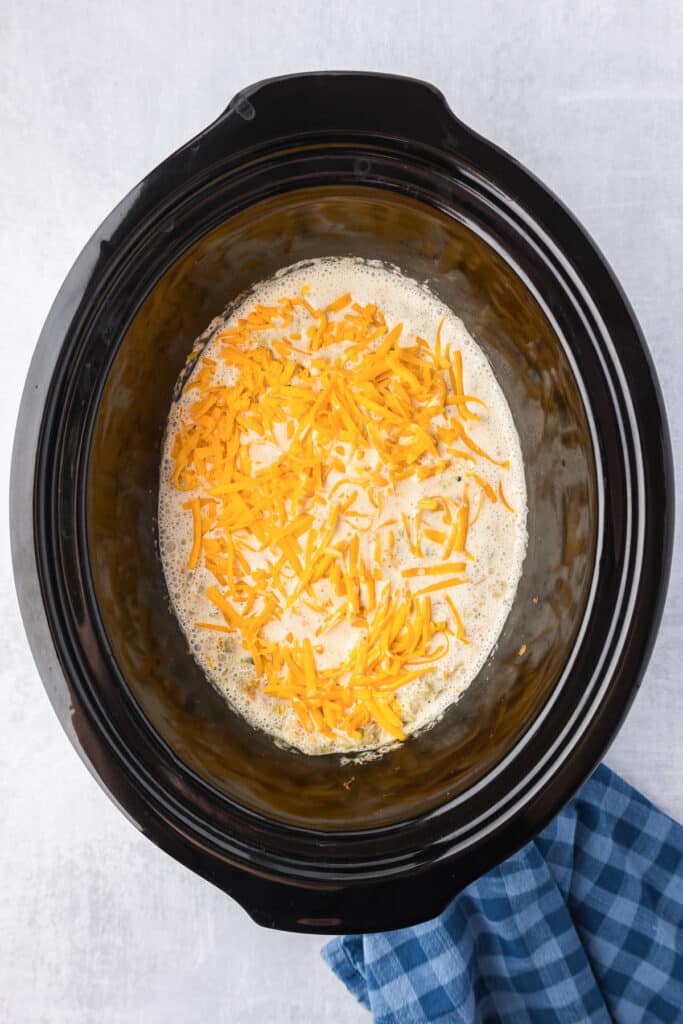 A crock pot with shredded cheese and cream inside for broccoli cheese soup.