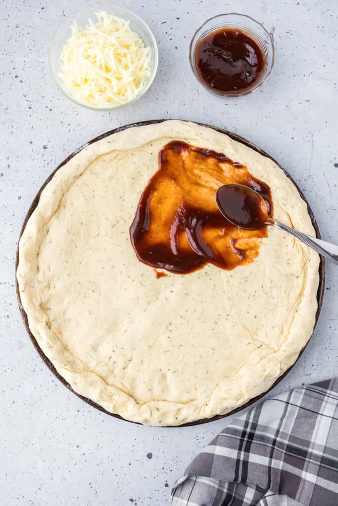 Pizza dough on a pan with bbq sauce being spread on top with a spoon.