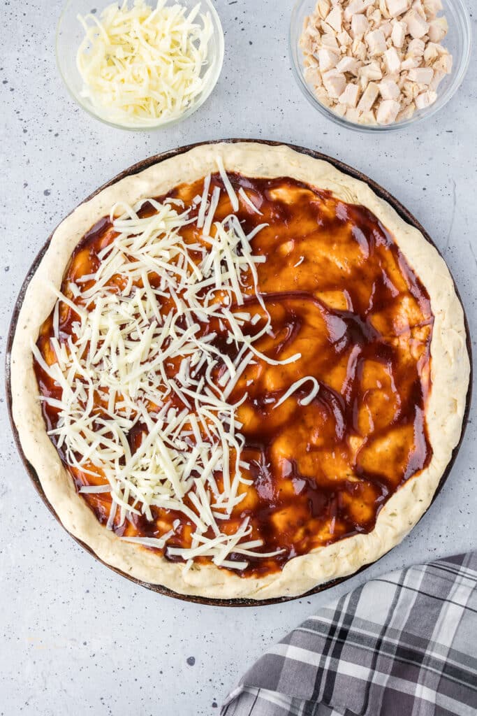 A pizza dough with bbq sauce and mozzarella cheese being spread on top.