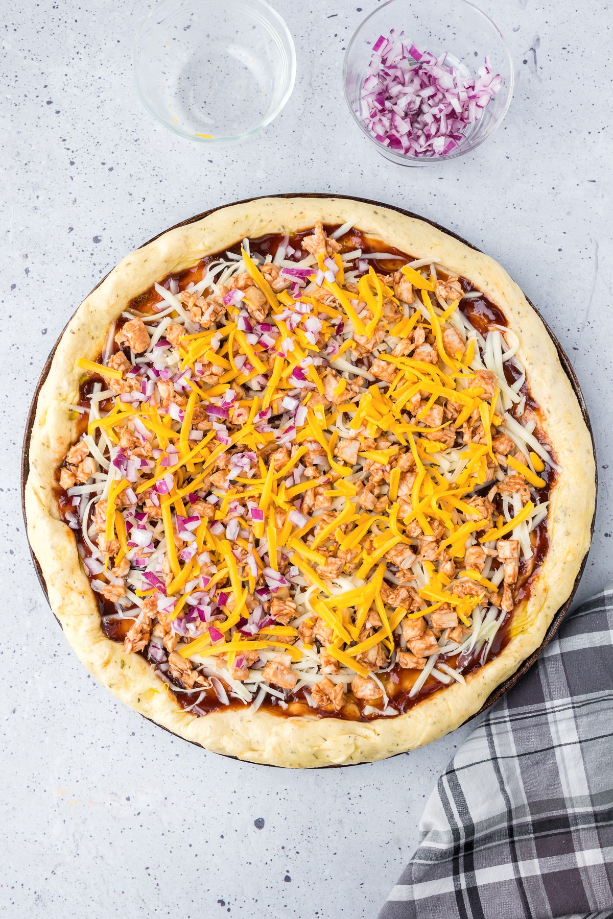 A bbw chicken pizza being sprinkles with cheddar cheese and chopped purple onion.