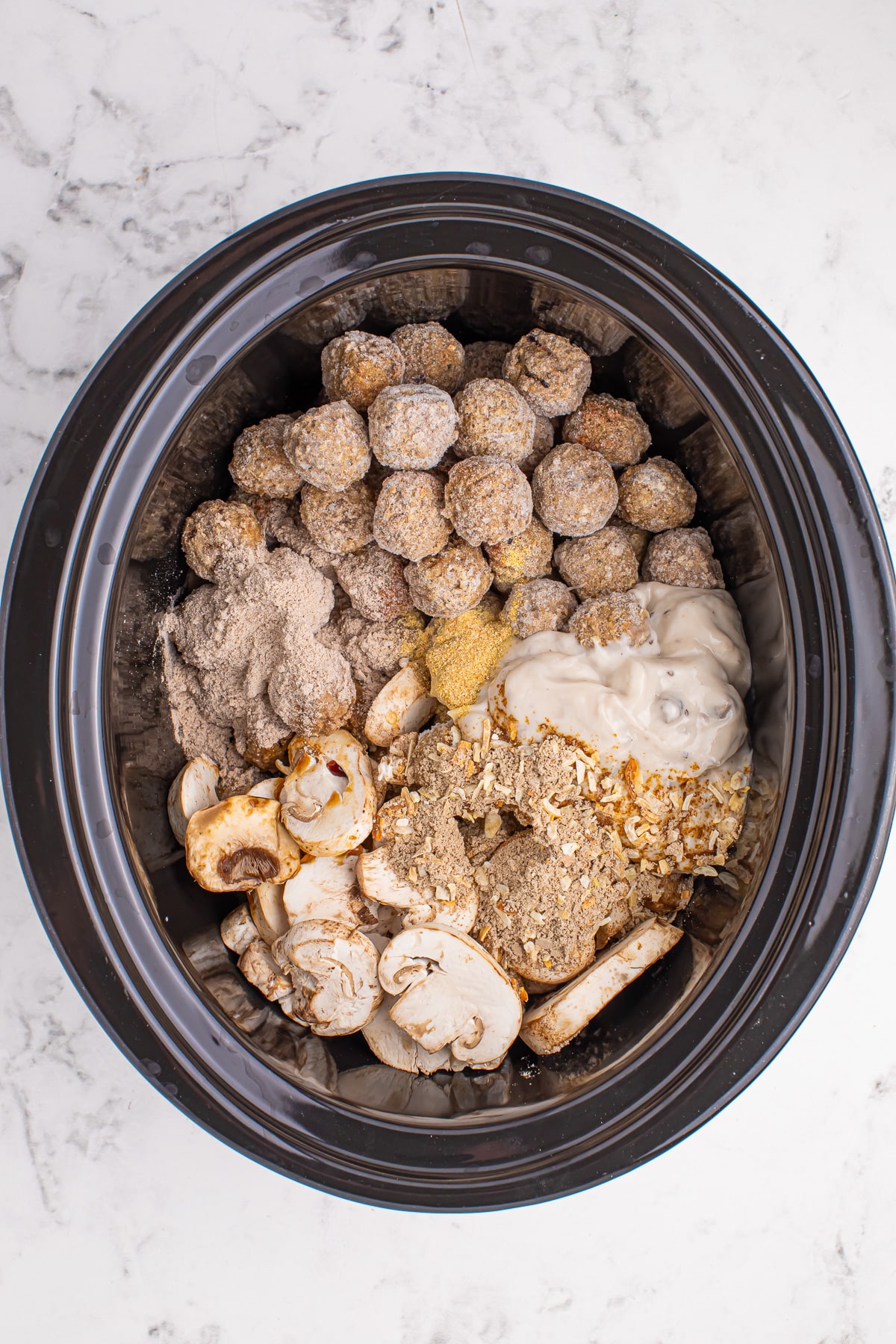A crock pot full of frozen meatballs, condensed soup, mushroom and spices for meatball stroganoff.