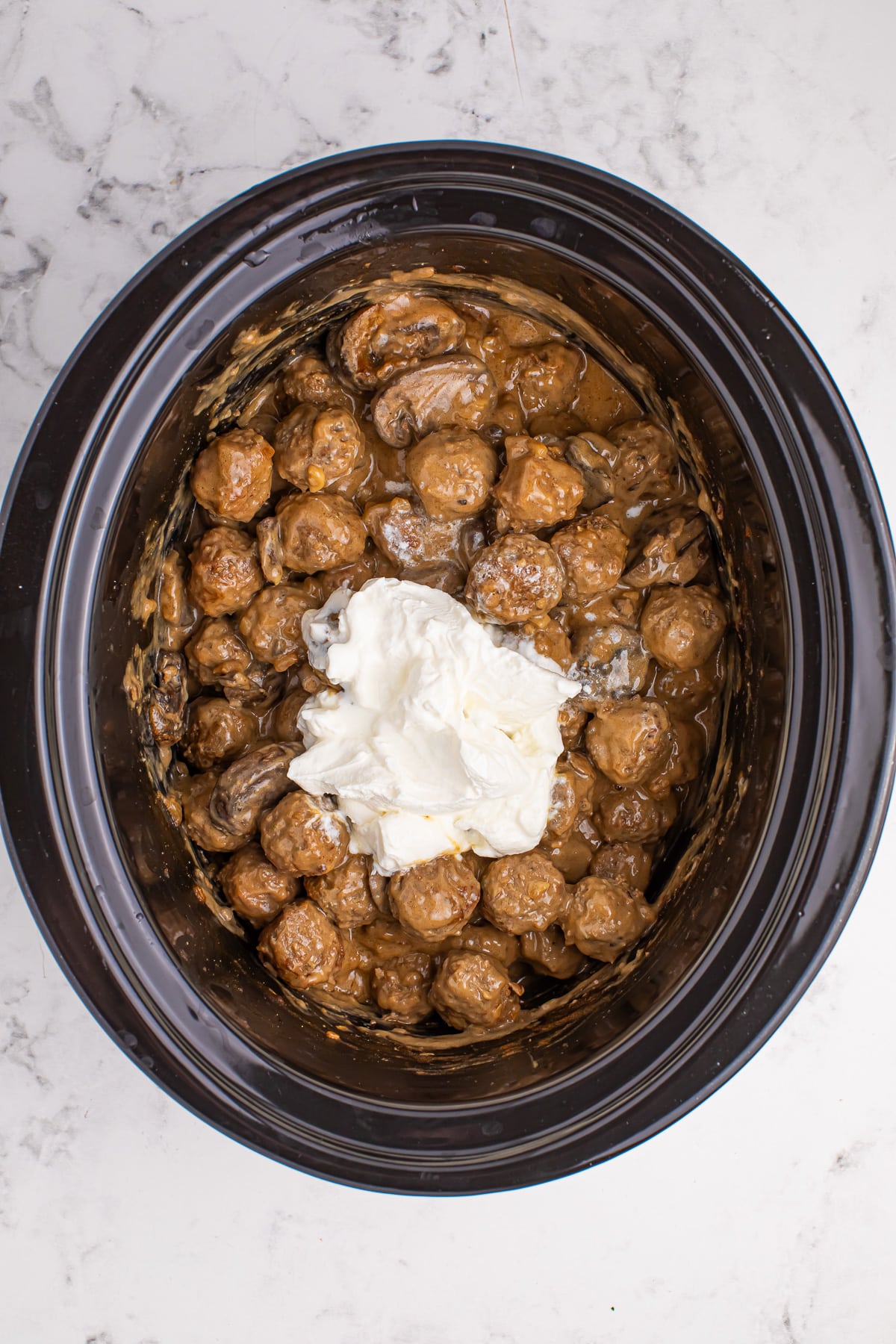 A crock pot filled with meatballs in sauce with a large amount of sour cream on top being mixed in.