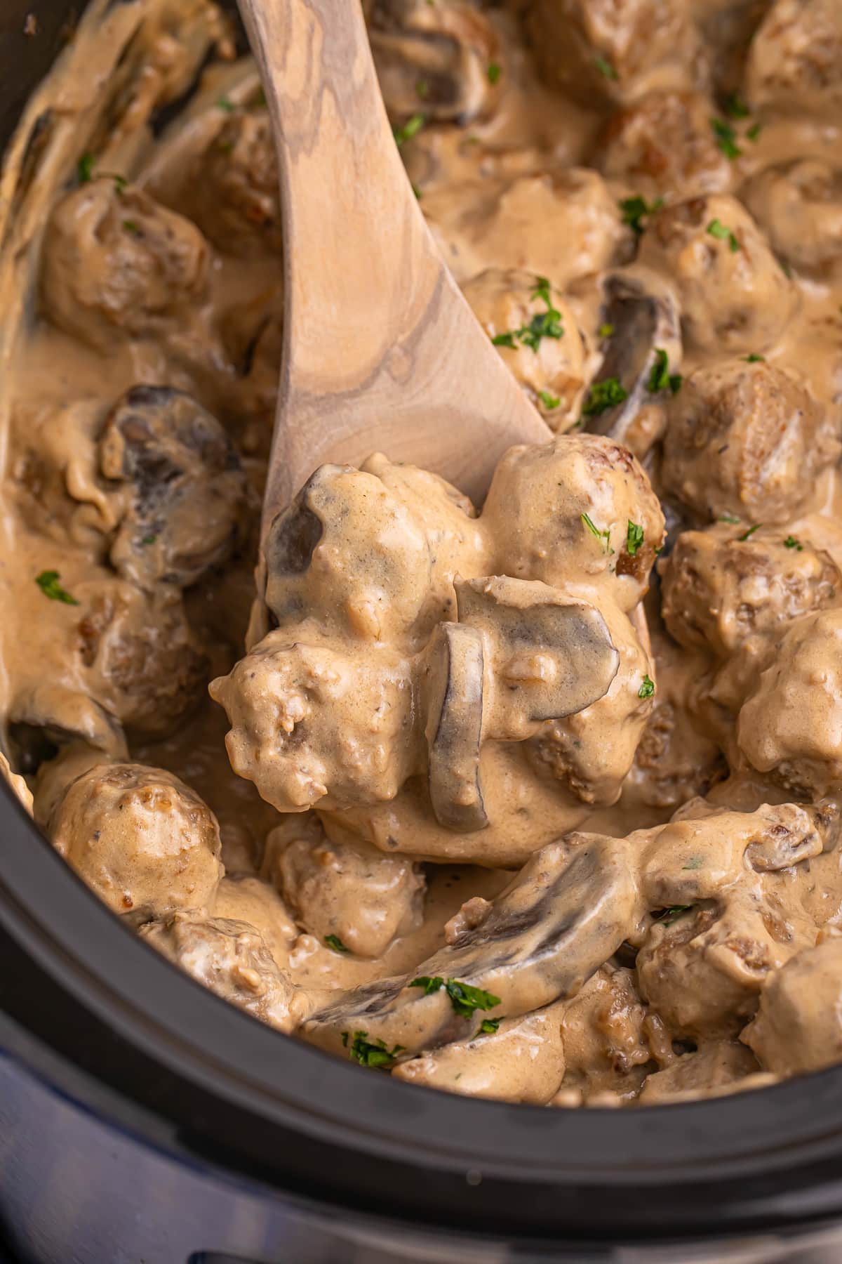A slow cooker filled with meatballs stroganoff being scooped by a spoon.
