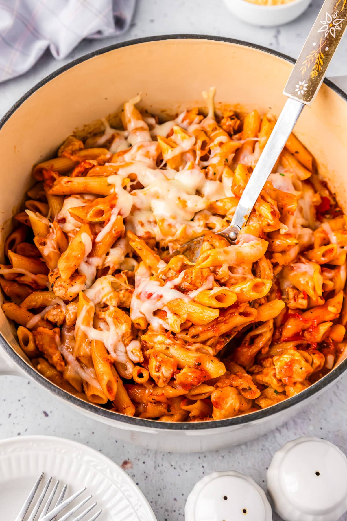 A pot full of chicken parmesan pasta with cheese on top and a large spoon scooping the penne pasta.