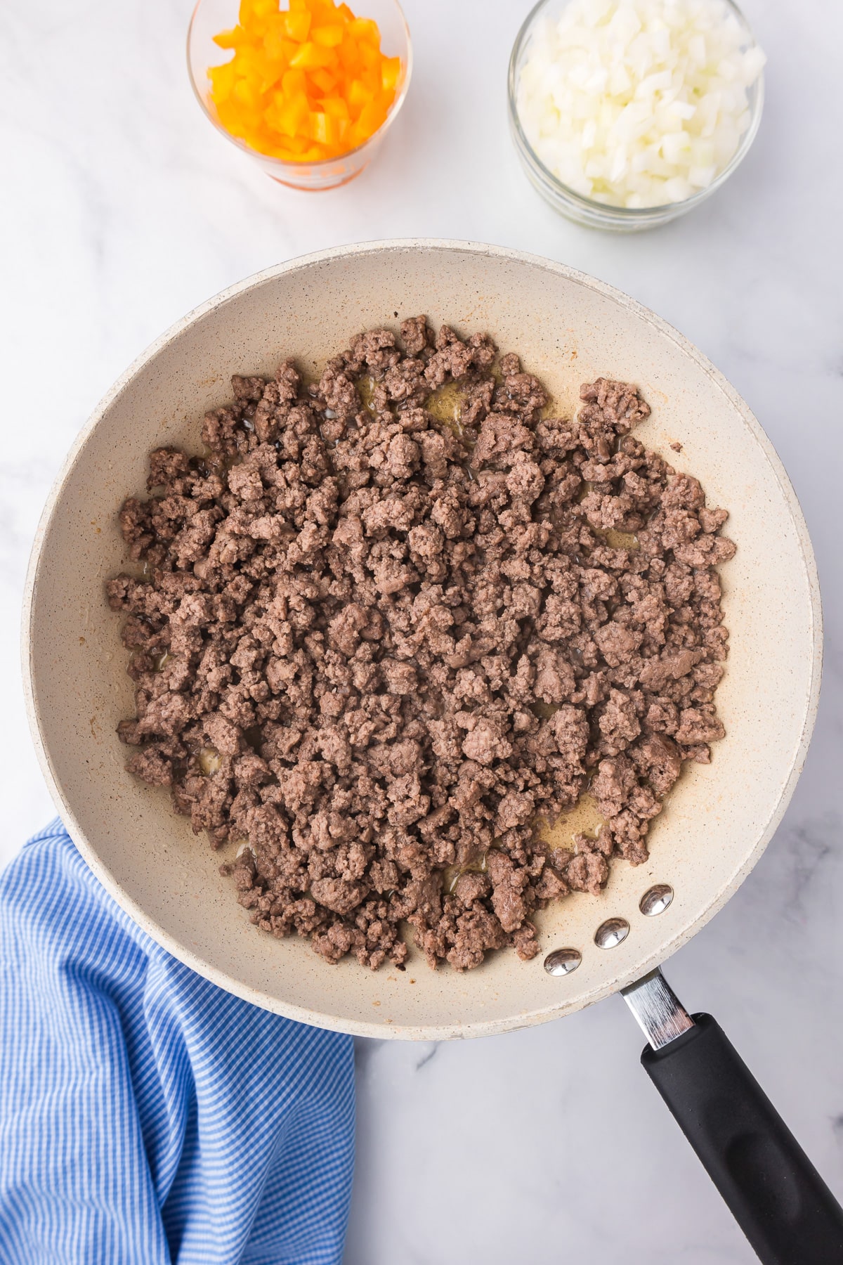 A frying pan filled with cooked ground beef for sloppy joes with bowls of diced bell pepper and diced onion nearby.