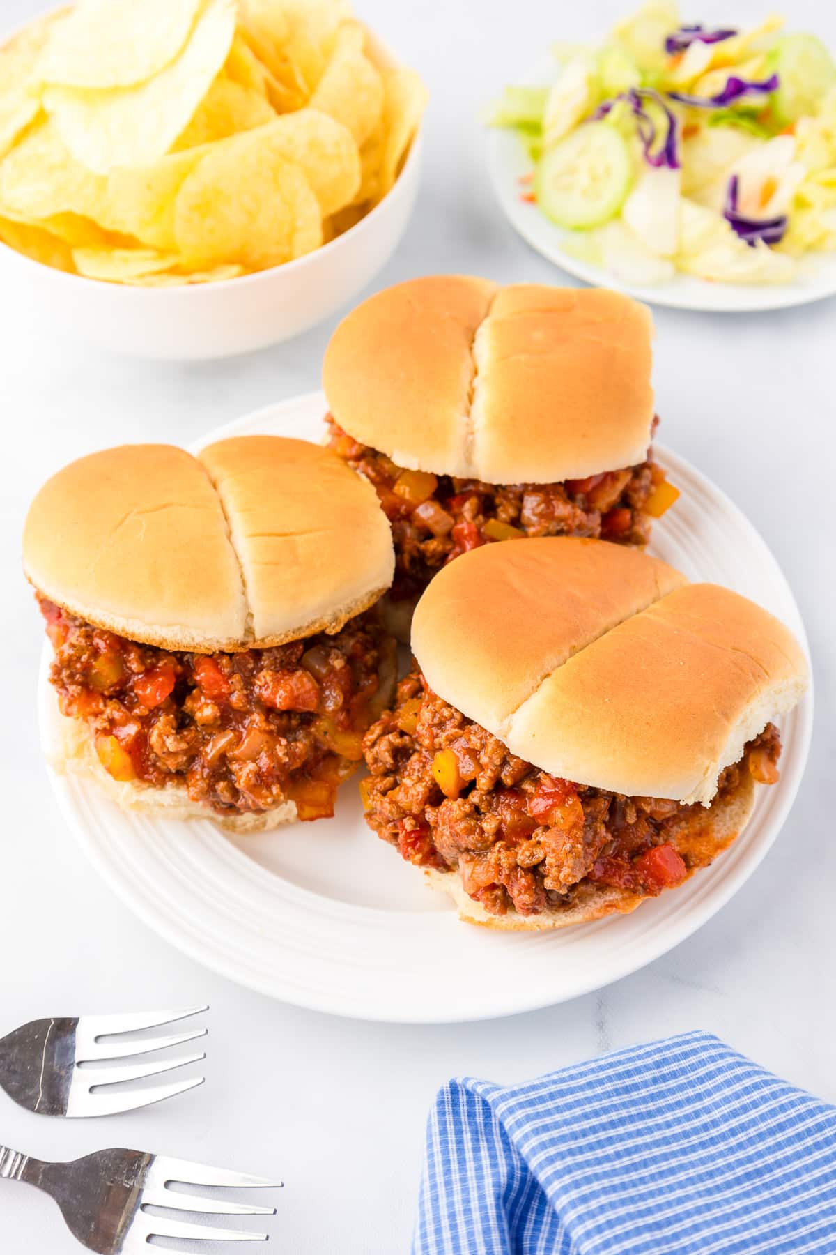 Three sloppy joes on a serving platter with chips and salad in bowls in the background.