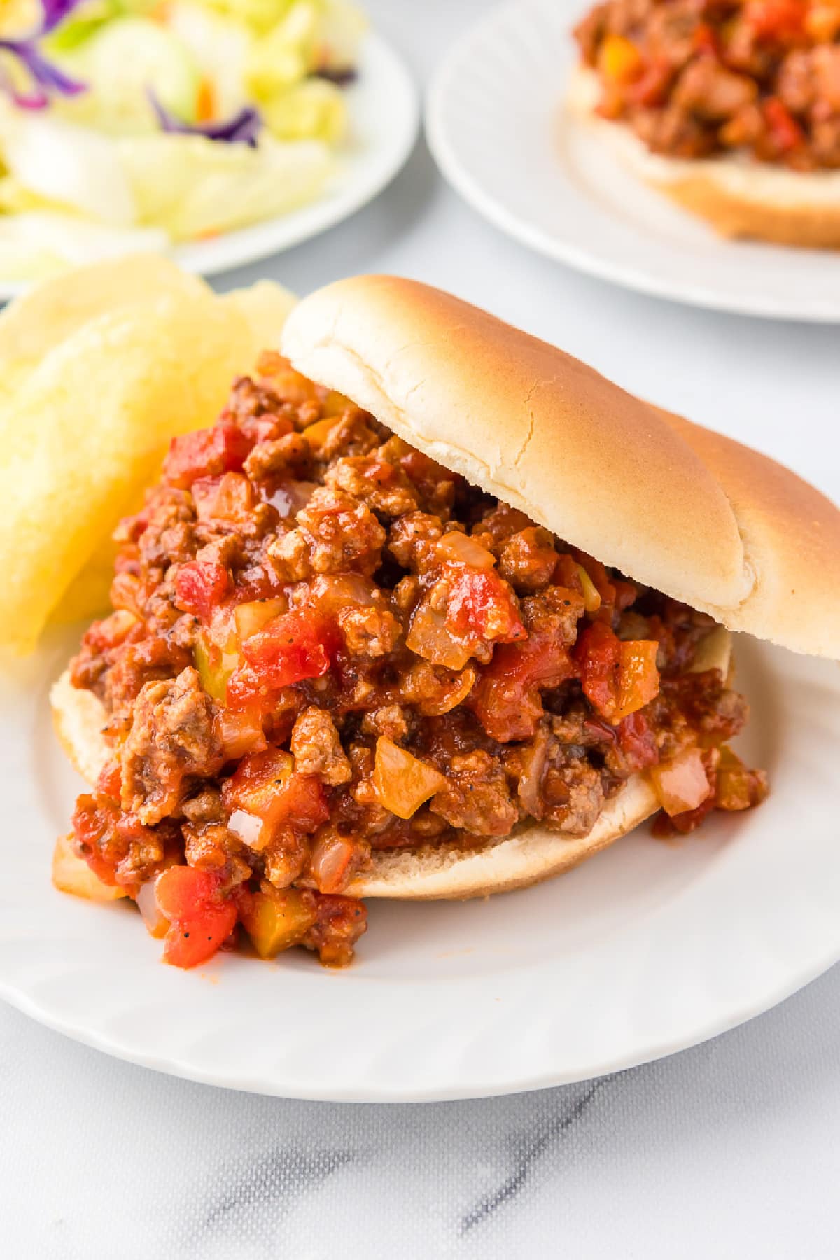 A sloppy joe on a plate next to chips with salad and a second sloppy joe sandwich in the background.