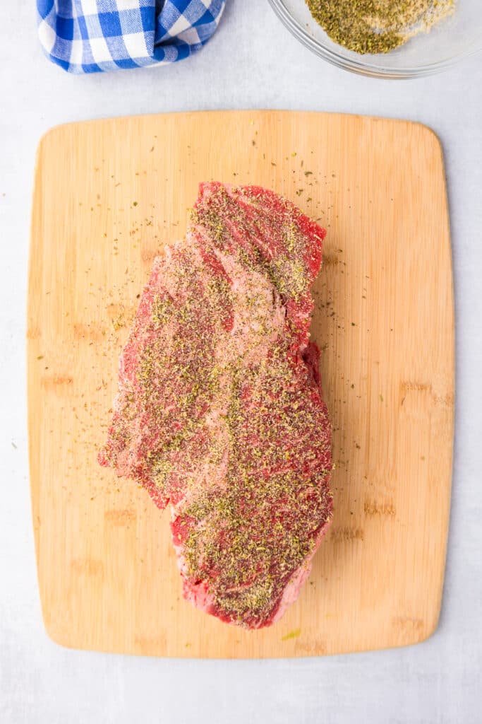 A piece of chuck roast beef on a cutting board with seasonings rubbed on it.