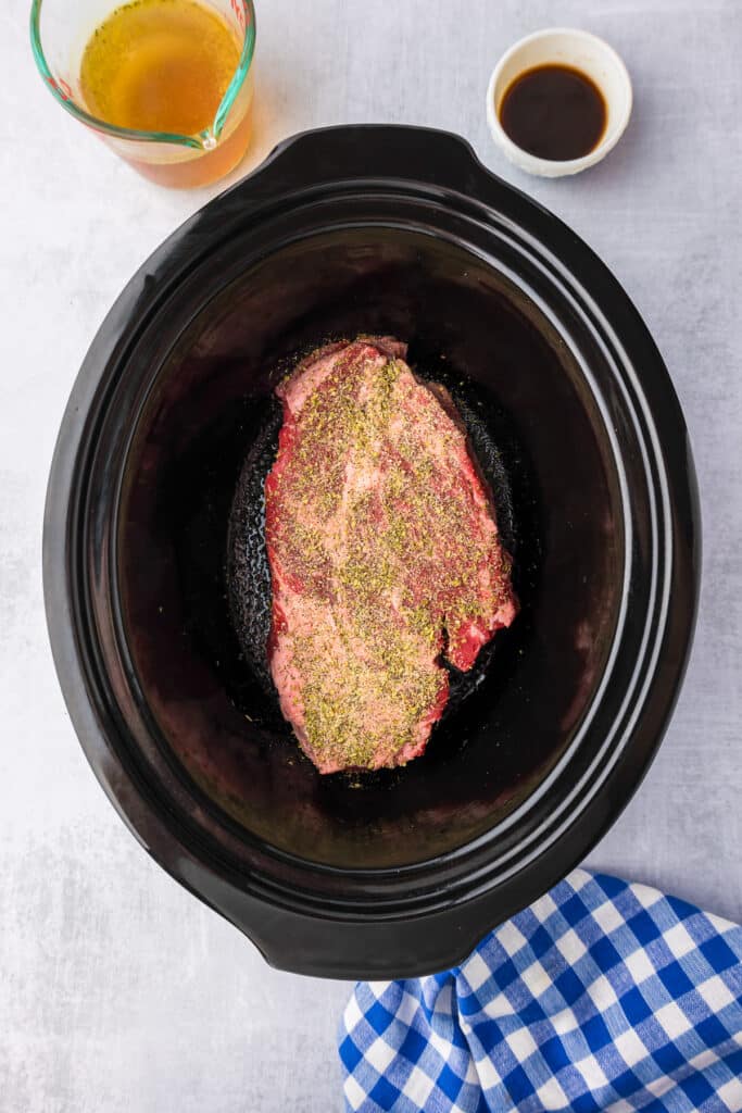 A crock pot base with a piece of seasoned beef roast inside, with broth and Worcestershire sauce in bowls nearby.