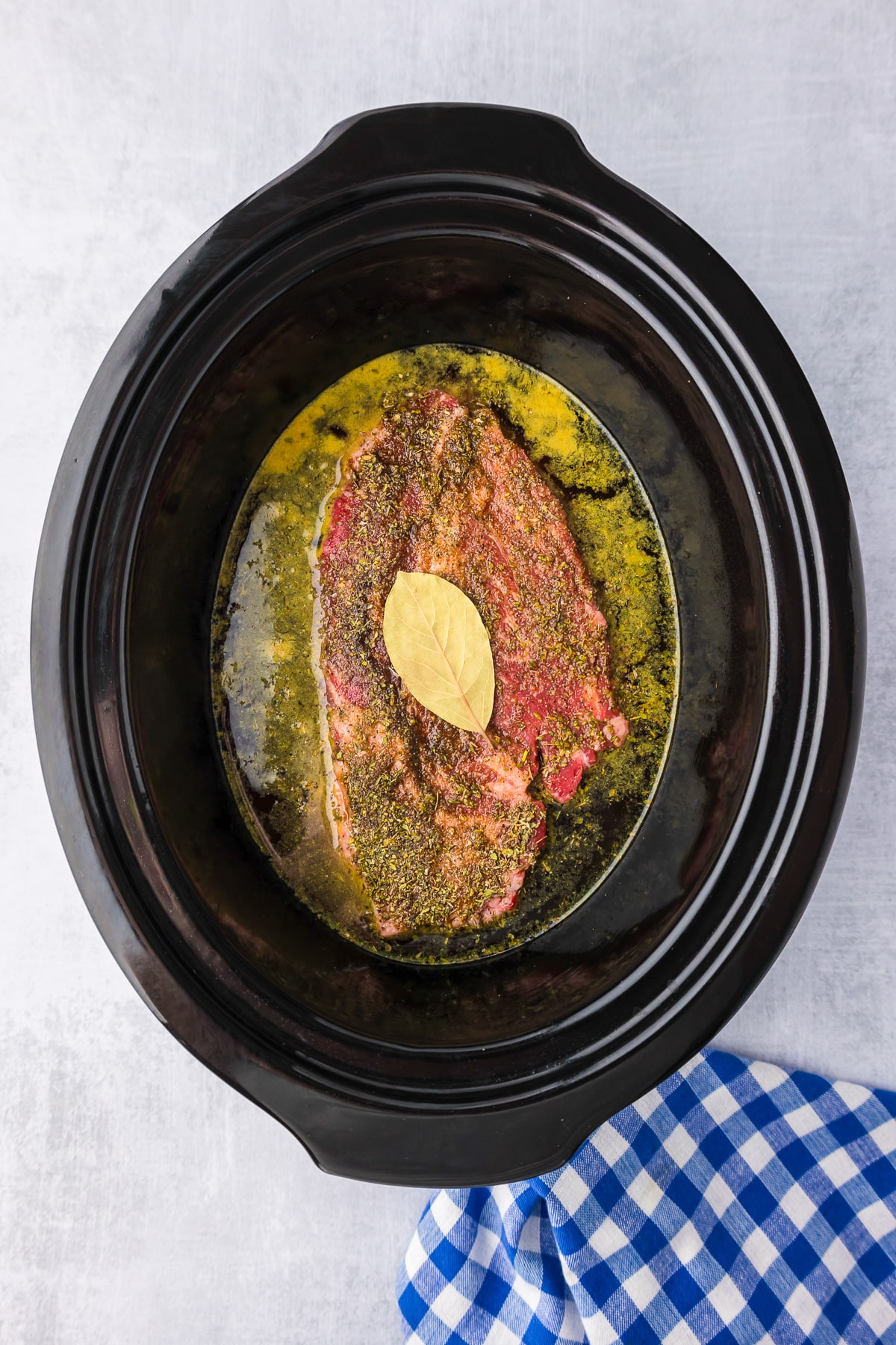 A crock pot base with a piece of seasoned beef topped with spices, broth and a bay leaf.
