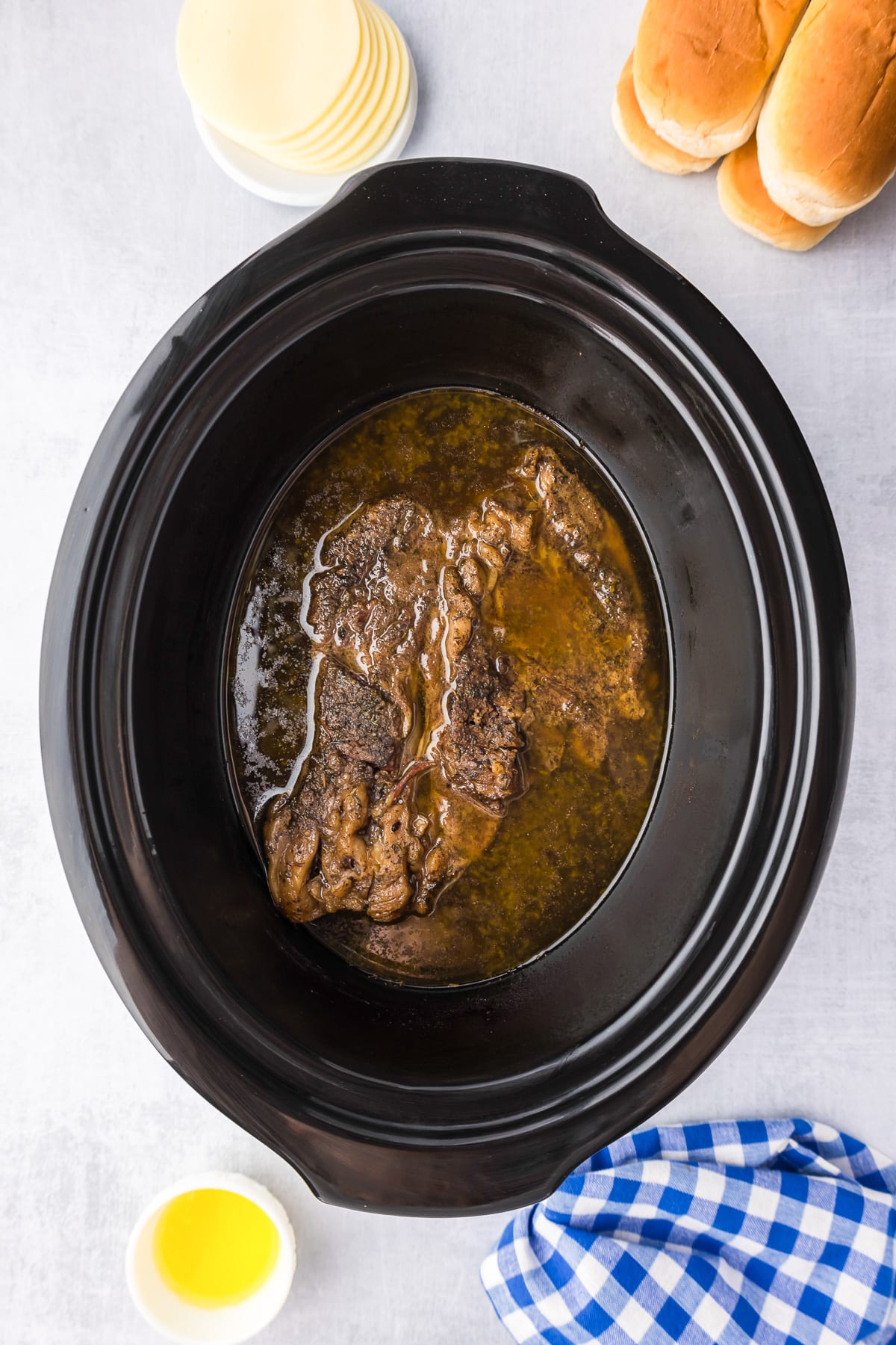 A crock pot filled with french dip sandwich beef and broth after cooking with rolls and cheese on the counter nearby.