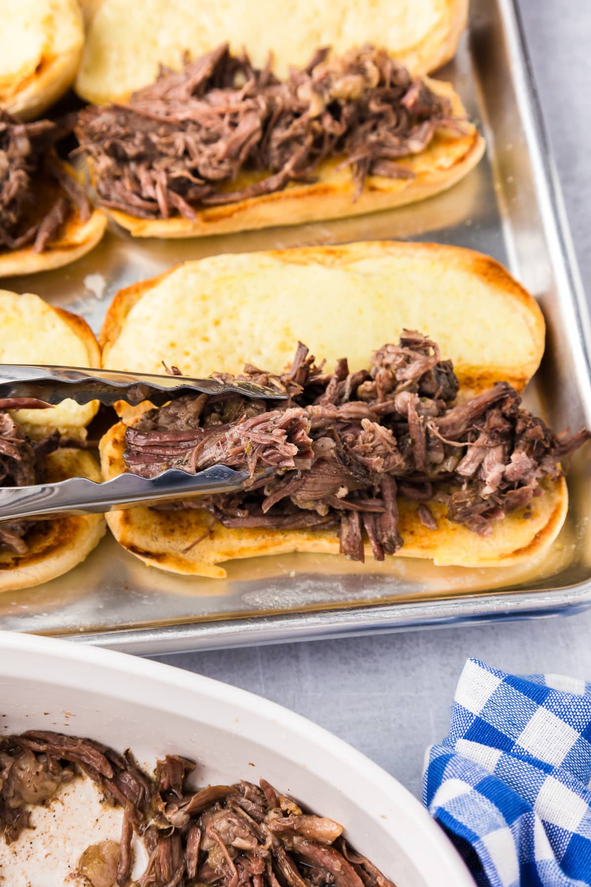 Placing shredded beef on toasted buns on a pan with tongs.