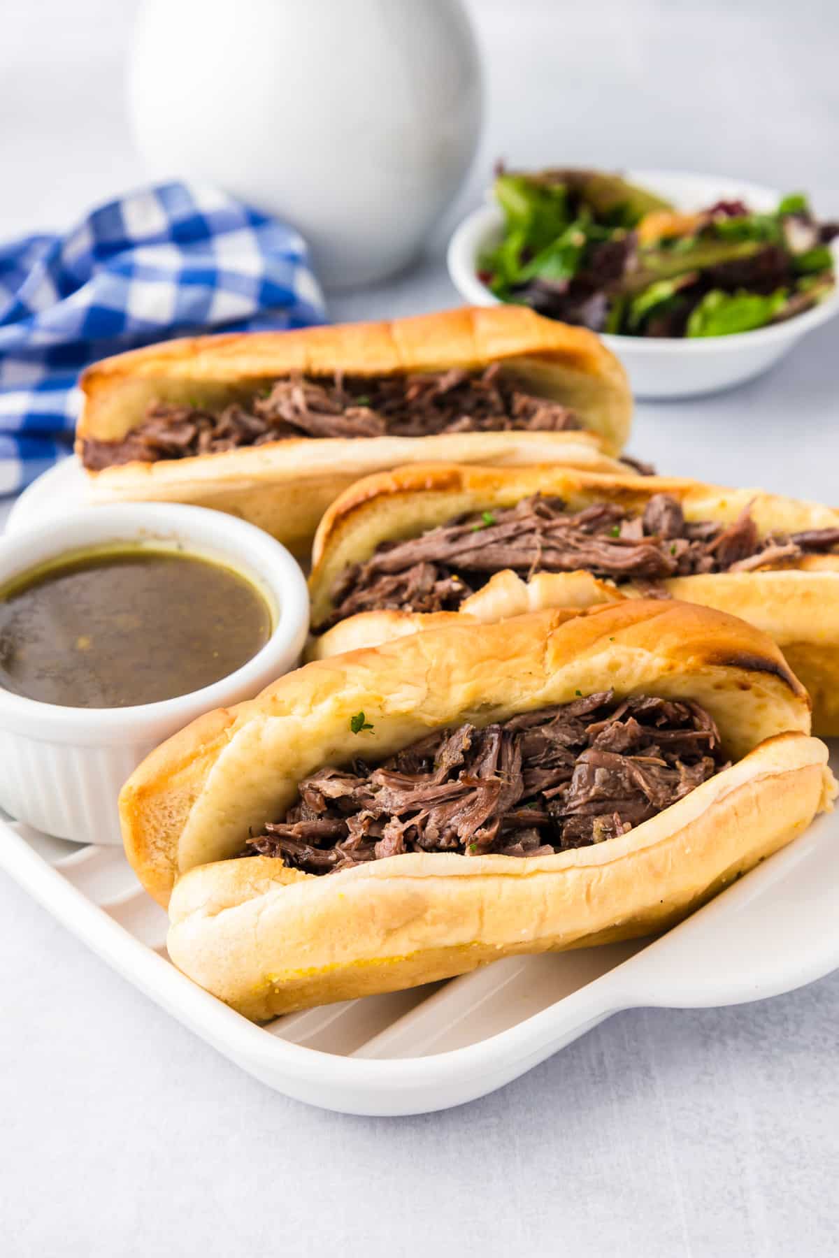A plater with three french dip sandwiches and a bowl of aus jus sauce next to them.