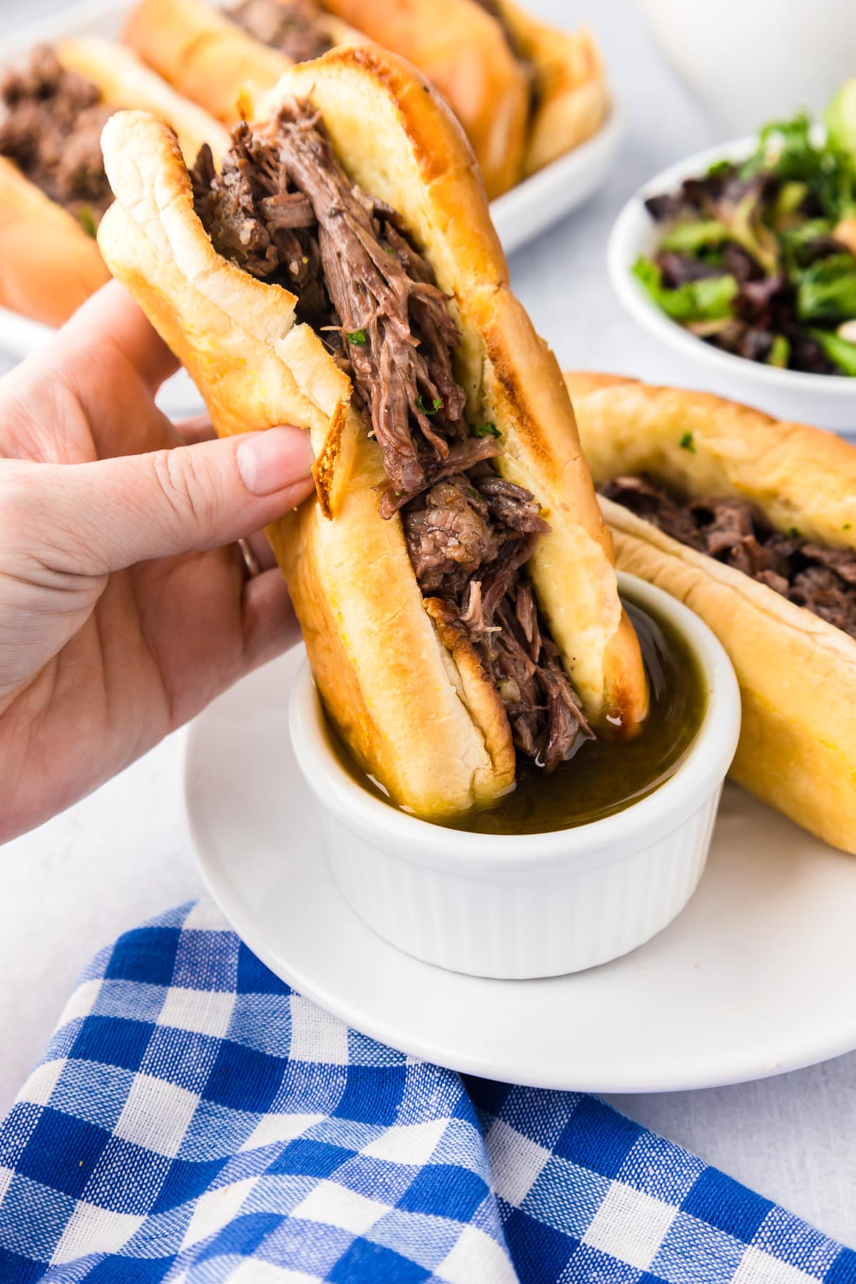 A person's hand holding a french dip sandwich and dipping it into aus jus sauce on a plate.