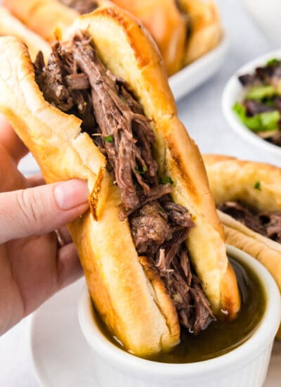 A person is holding a French dip sandwich with beef dipping it into an aus jus sauce.