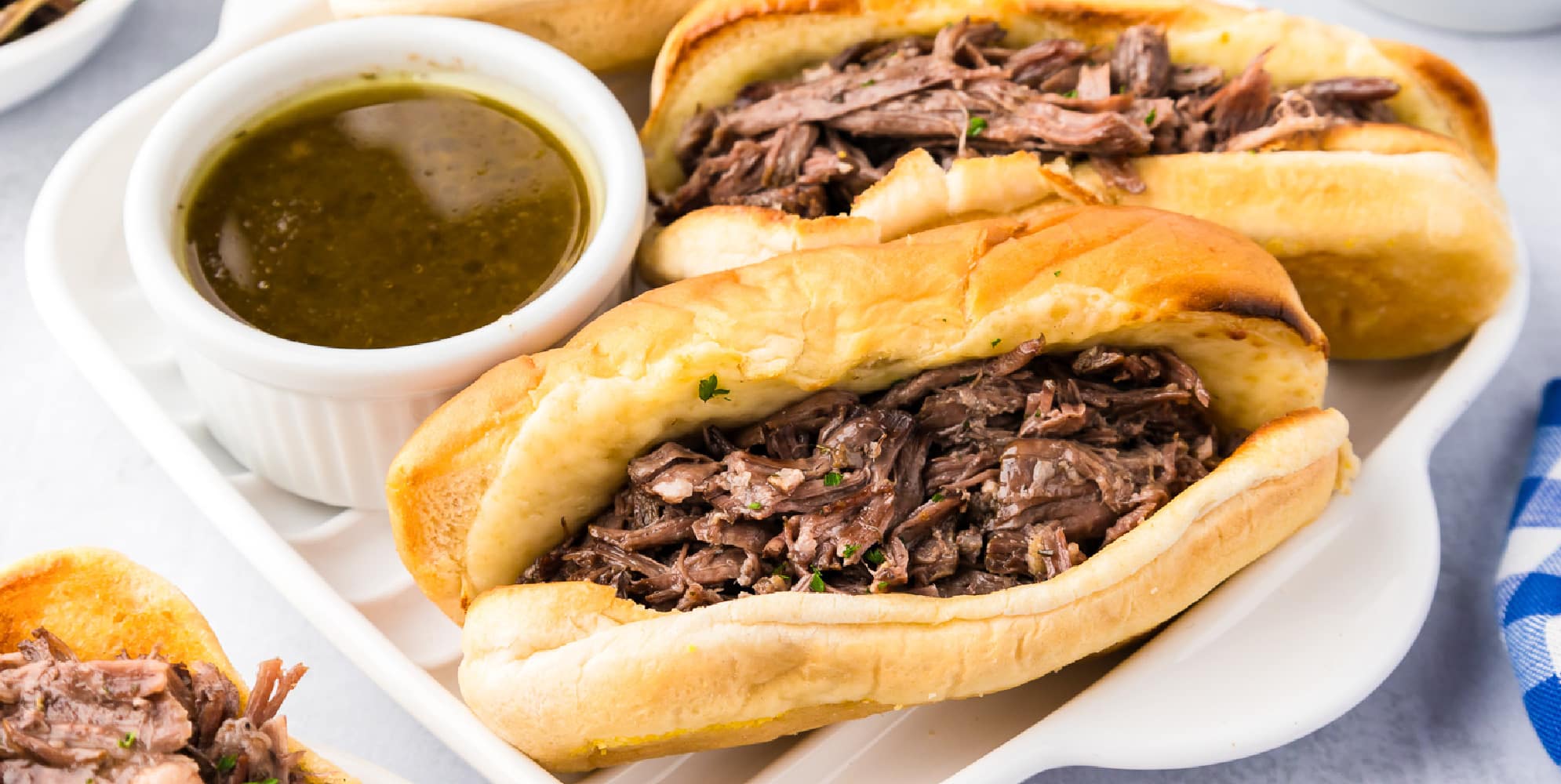 A platter with several French dip sandwiches and aus jus dipping sauce in a bowl.