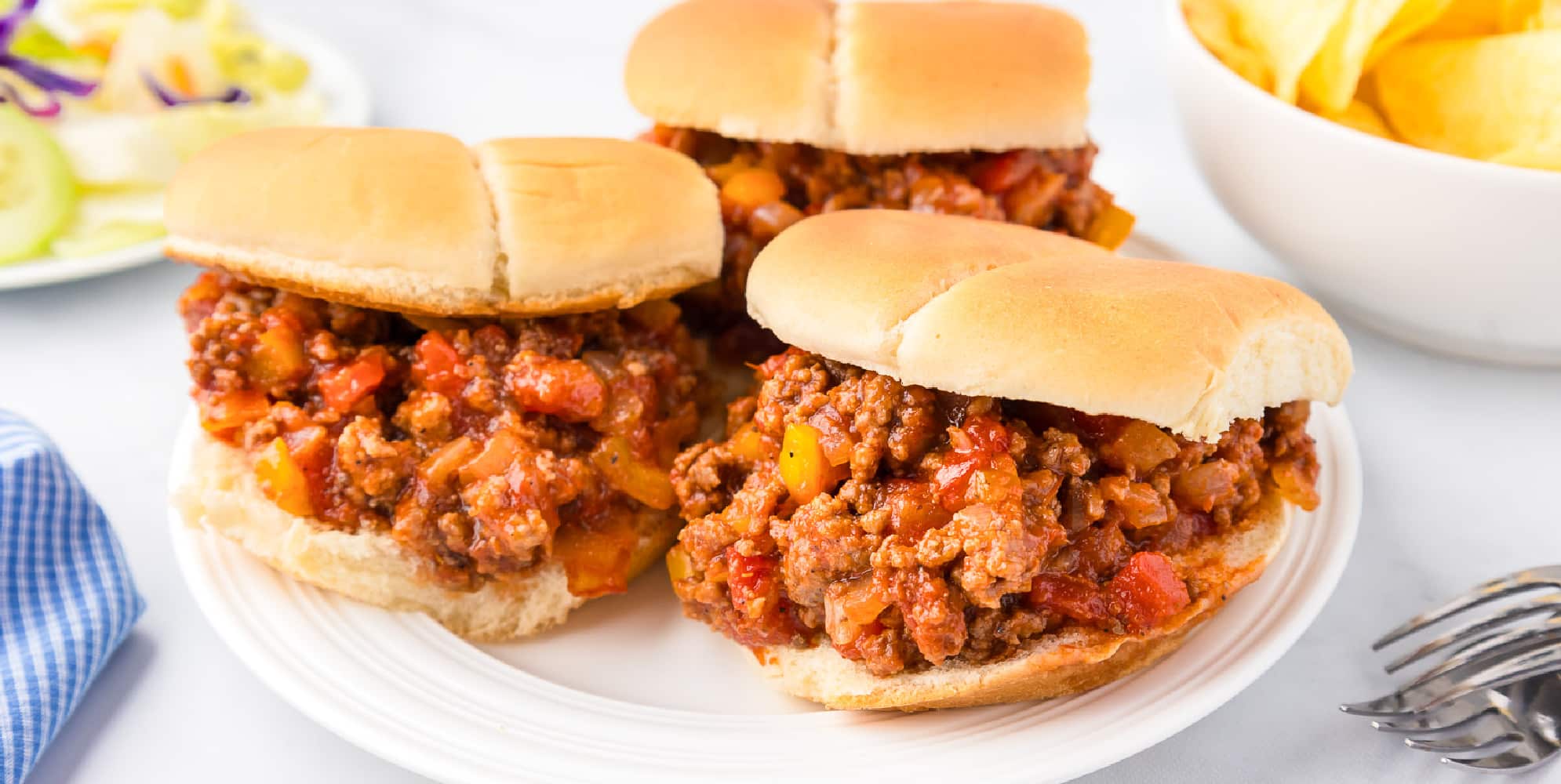 Wide view of three sloppy joes on a plate with chips.