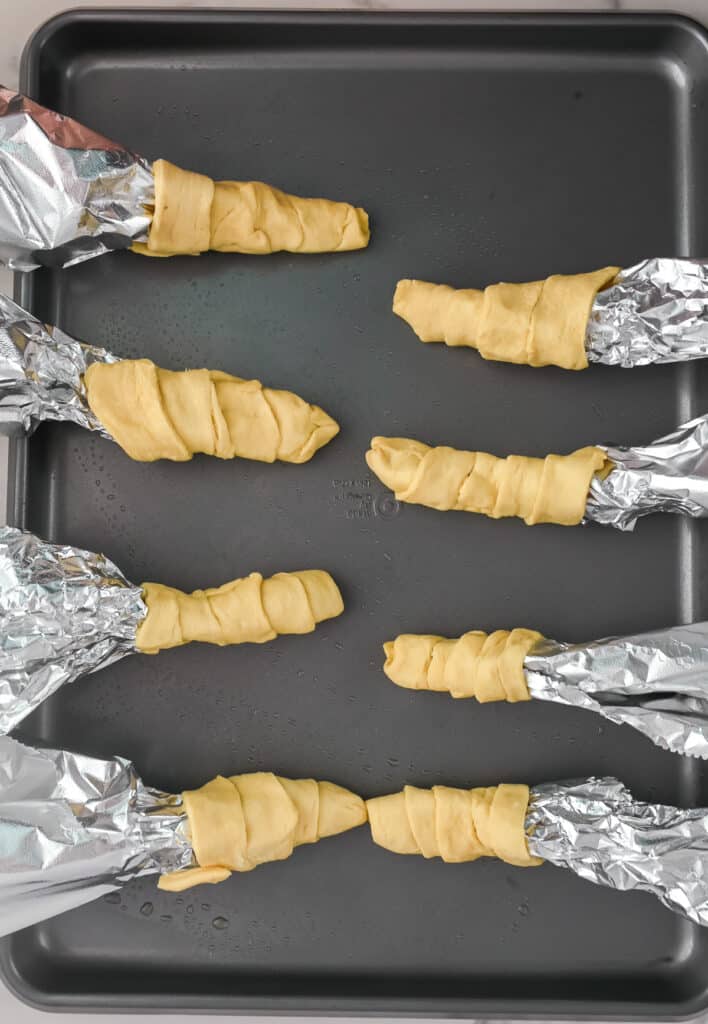 A baking sheet with crescent roll dough wrapped around pieces of aluminum foil.