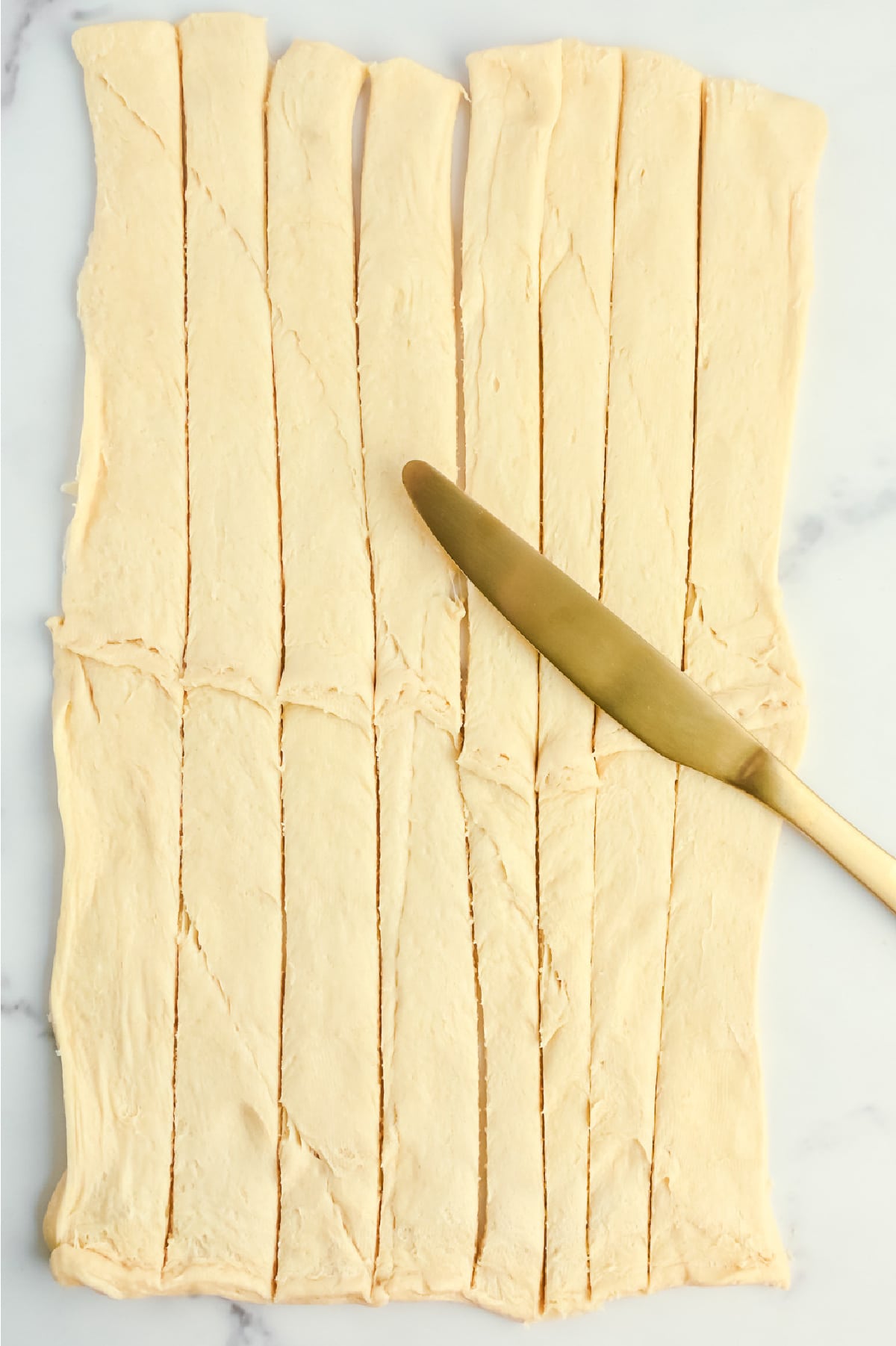 A piece of crescent roll dough on a counter cut into long strips with a knife on top.