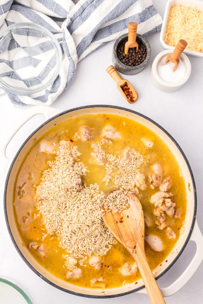 Chicken, broth and uncooked rice in a pan with a wooden spoon to make creamy chicken and rice.