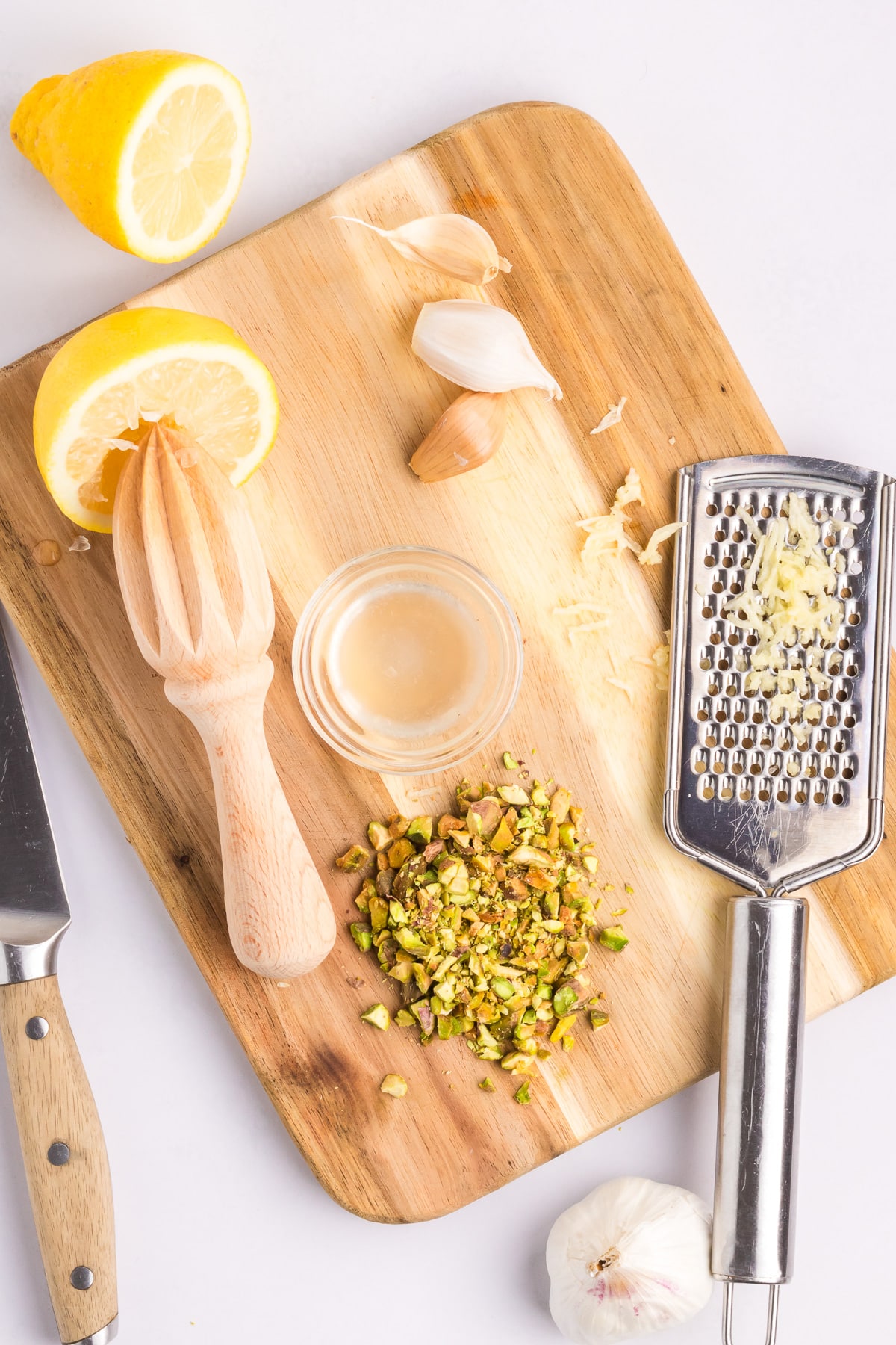 A cutting board with chopped pistachips, grated parmesan cheese, garlic cloves and lemon halves.