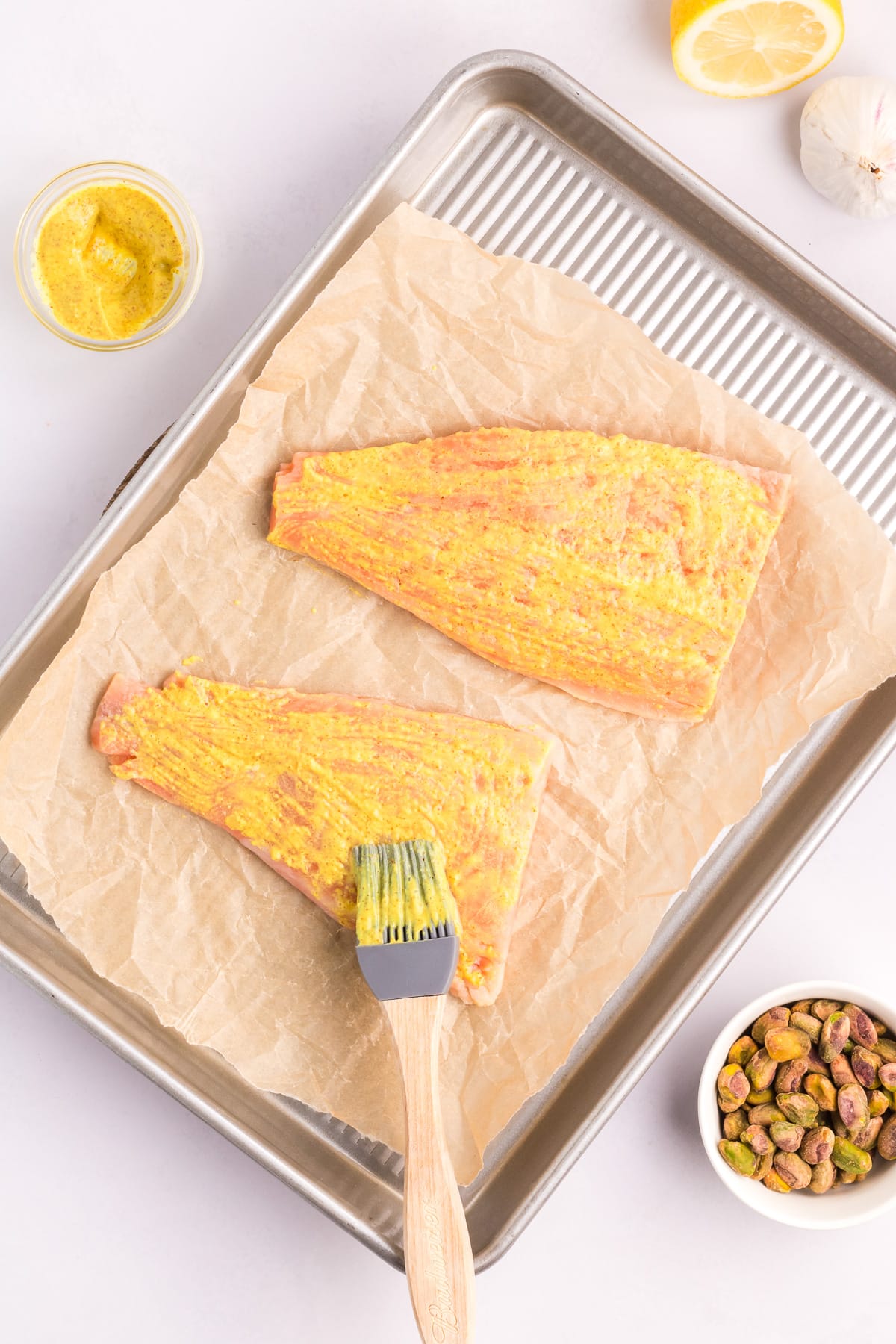 A silicone brush spreading Dijon mustard on two salmon filets on a baking sheet.