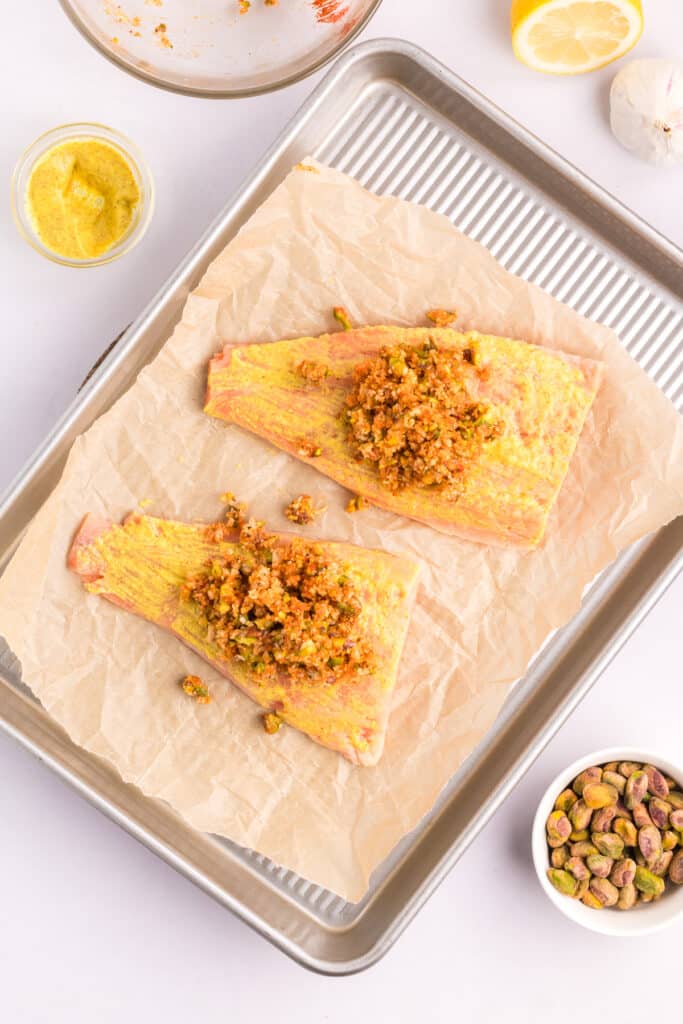 Two salmon fillets on a baking sheet coated in Dijon mustard with pistachio crust mixture being added on top.