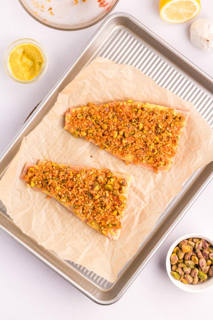 Two salmon fillets on a baking sheet coated in Dijon mustard with the pistachio crust fully coating the fillets.