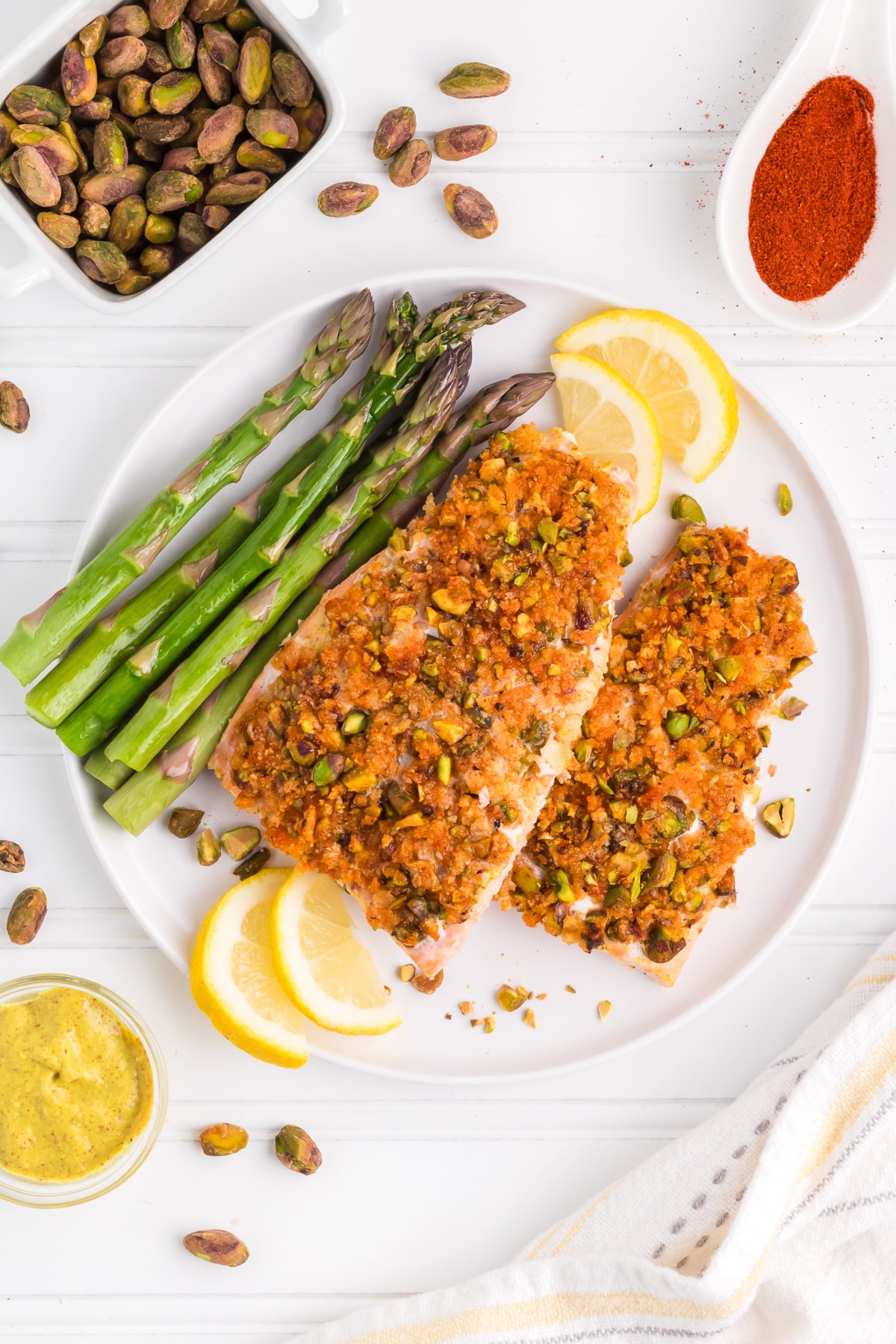 A plate with pistachio crusted salmon filets, asparagus, and lemon slices on a table.