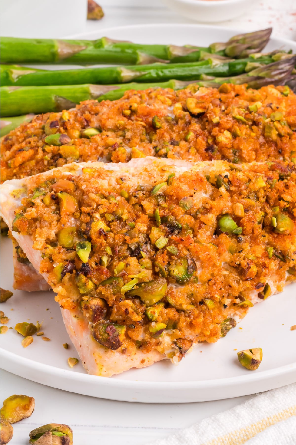 Two crusted pistachio salmon fillets on a plate from the side with asparagus next to it.