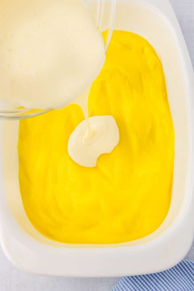 A cream cheese mixture being poured over a lemon pie filling mixture in a pan.