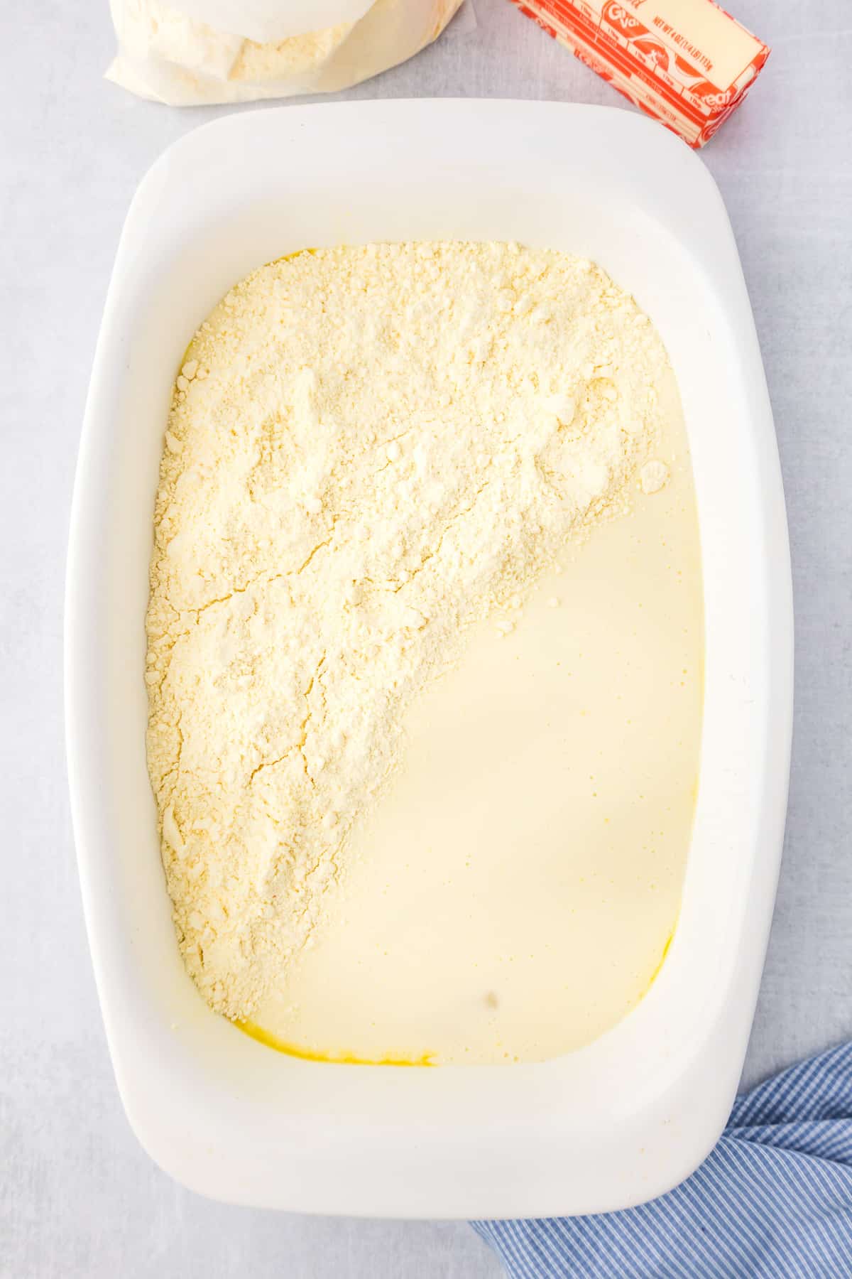A white pan filled with lemon and cream cheese layers being topped with dry yellow cake mix.