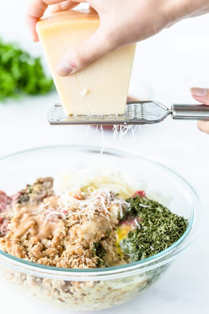 A person grating parmesan cheese into a bowl of meatloaf ingredients.