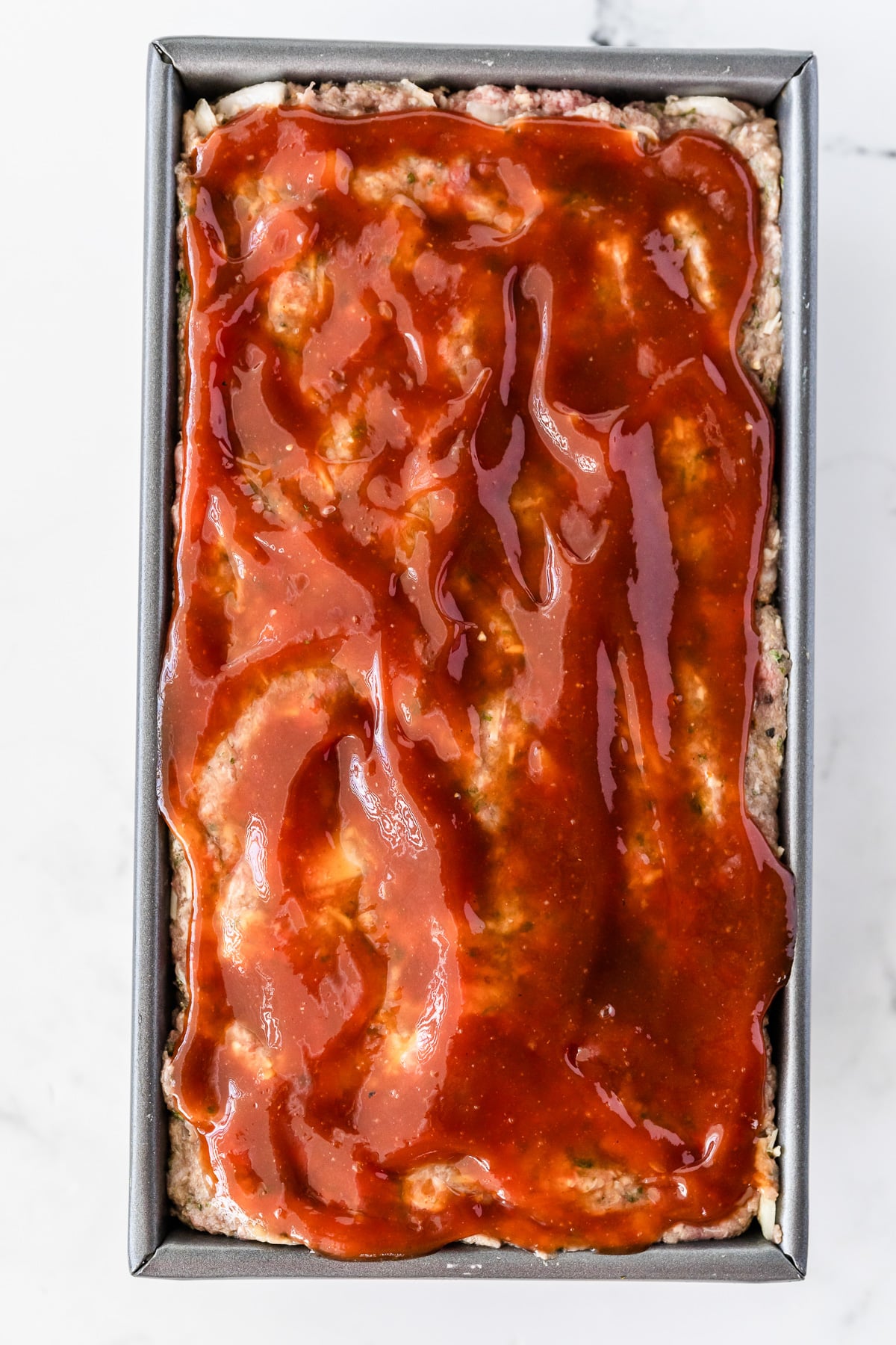 A raw meatloaf in a metal pan with a red sauce on top.