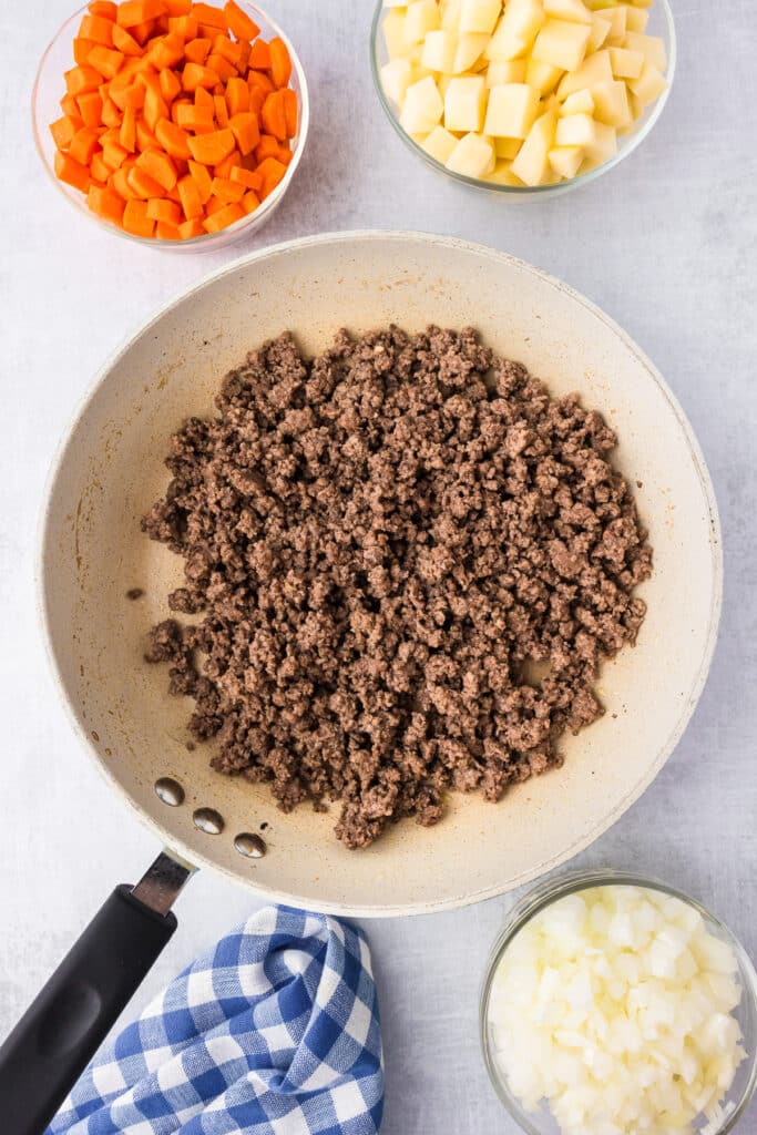 Ground beef in a frying pan after browning with chopped carrots potatoes and onions in bowls nearby..
