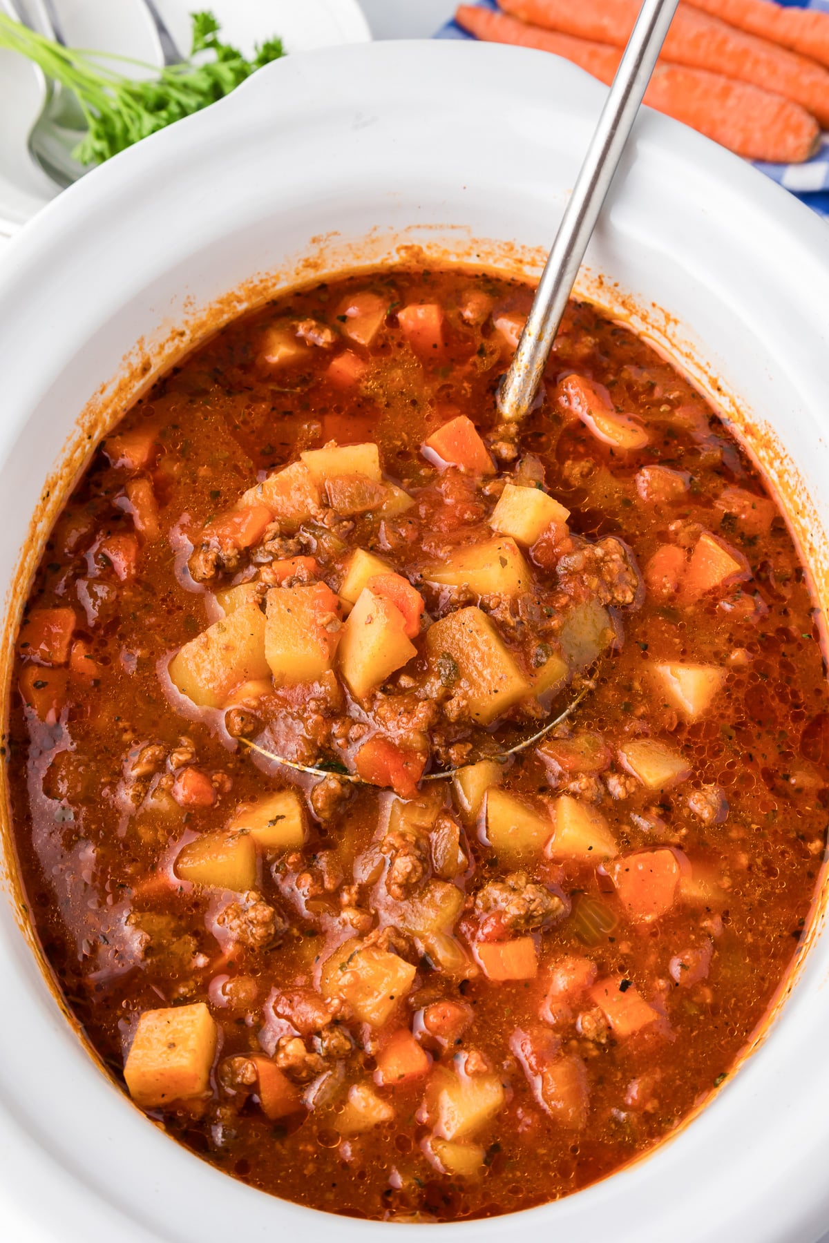 A bowl of hamburger stew with carrots and potatoes in a white slow cooker base being scooped from above.