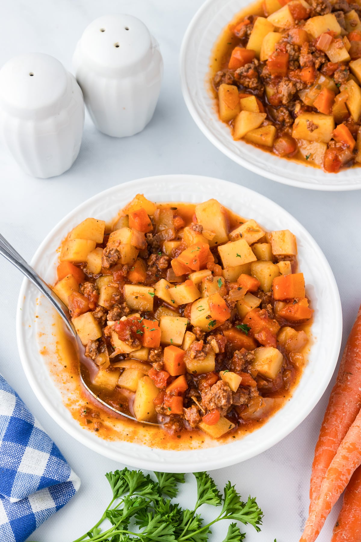 Two bowls of thick hamburger stew with carrots and potatoes in a bowl with a spoon scooping.