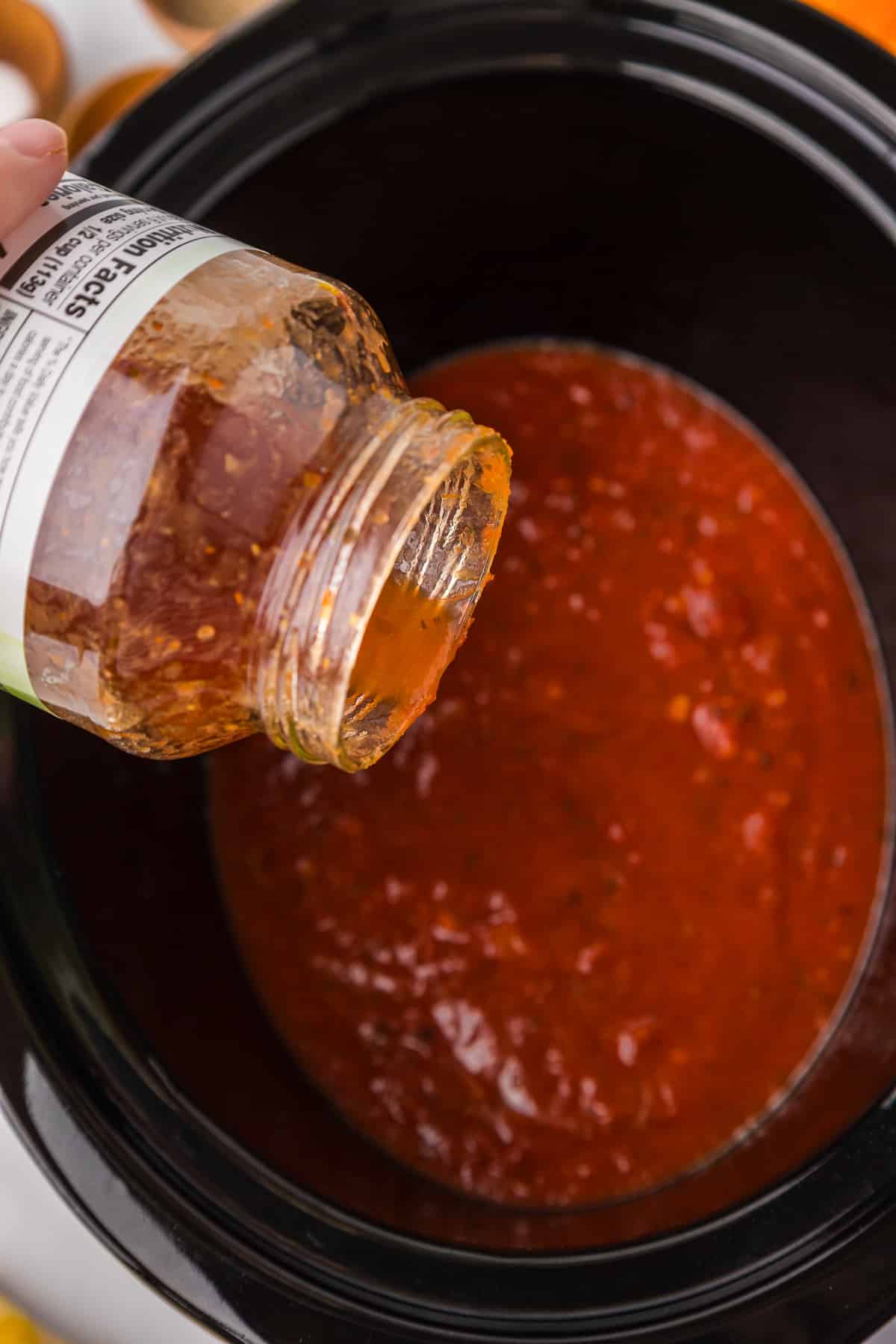 A person pouring marinara sauce into a slow cooker from a jar.