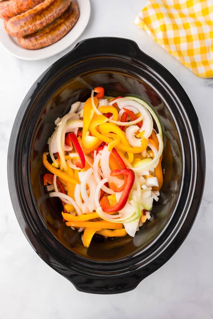 A slow cooker full of peppers and onions sliced into strips with browned sausages on a plate nearby.