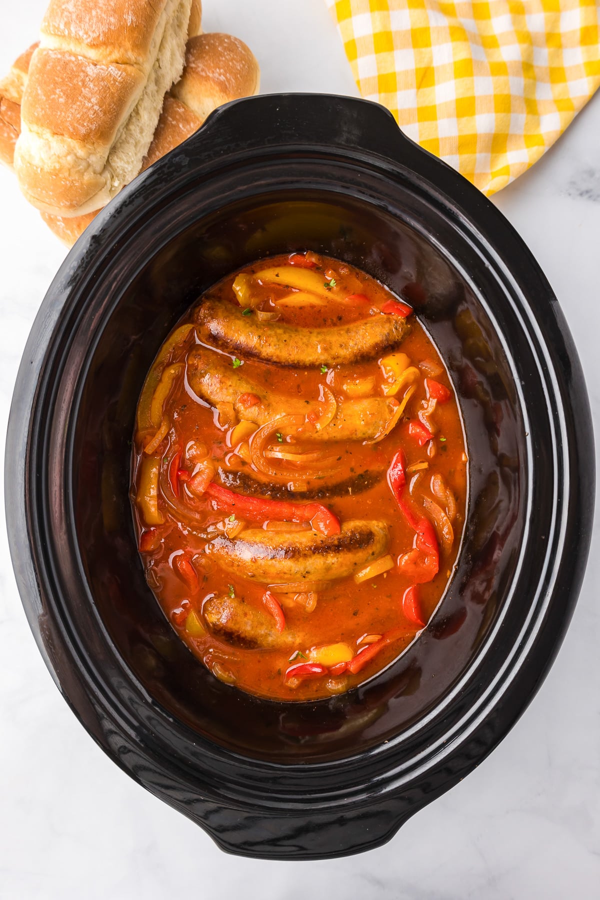A crock pot full of Italian sausages and peppers in a red marinara sauce with rolls nearby.