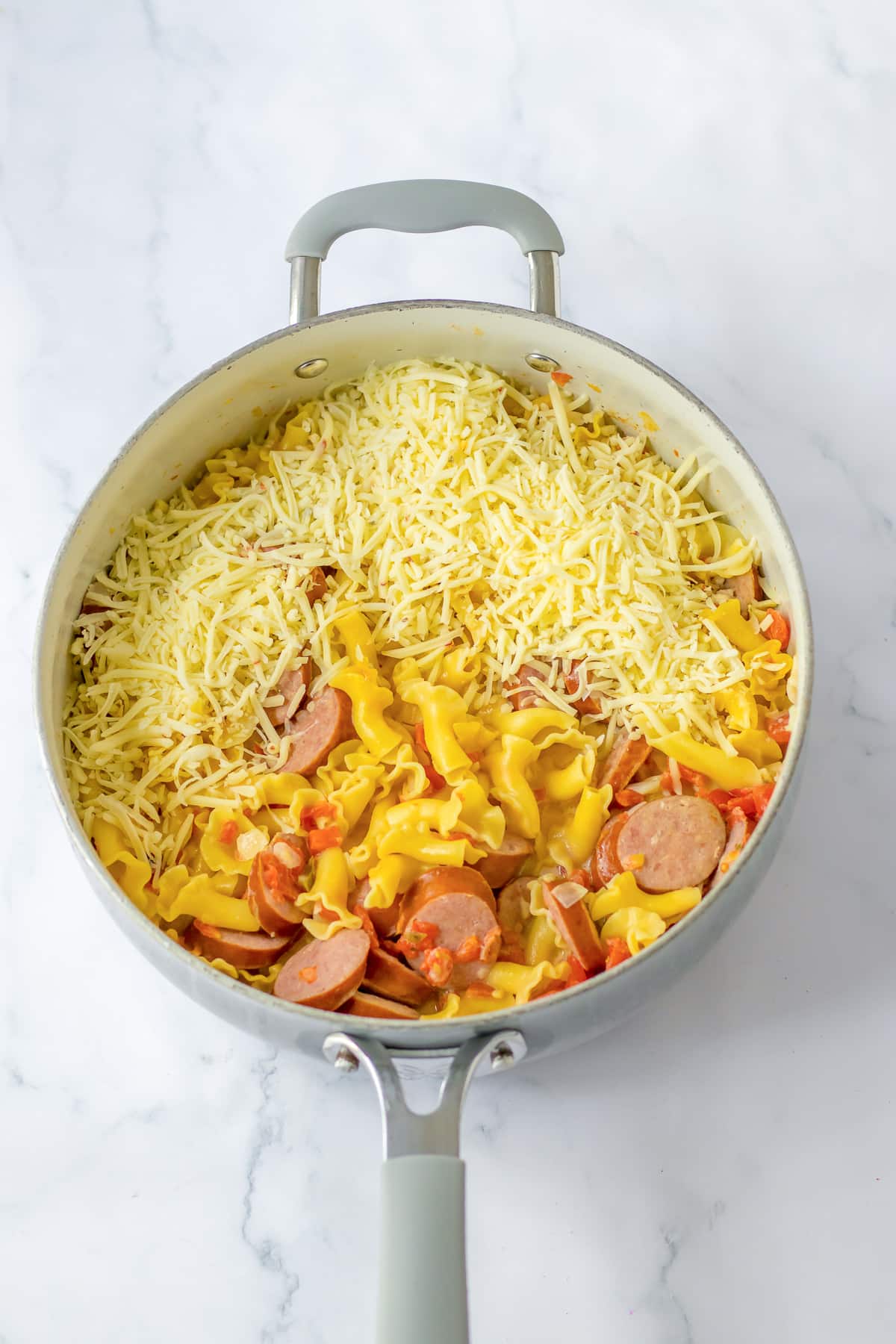A skillet full of smoked sausage and pasta in a creamy sauce being covered with shredded cheese.