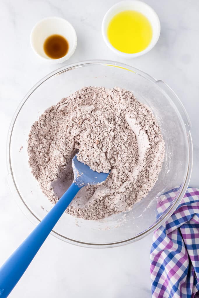 A spatula mixing together powdered sugar and cocoa with vanilla and egg whites in bowls nearby.
