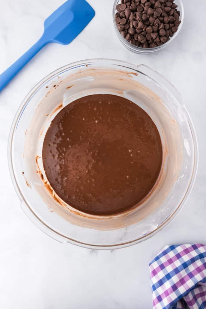 A liquidy flourless chocolate brownie cookie batter in a large mixing bowl with a bowl of chocolate chips nearby.